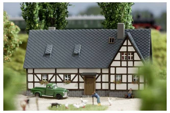 Miniaturmodell eines Fachwerkhauses mit einem grünen Pickup-Truck und winzigen Figuren davor, in einer landschaftlich gestalteten Szene.