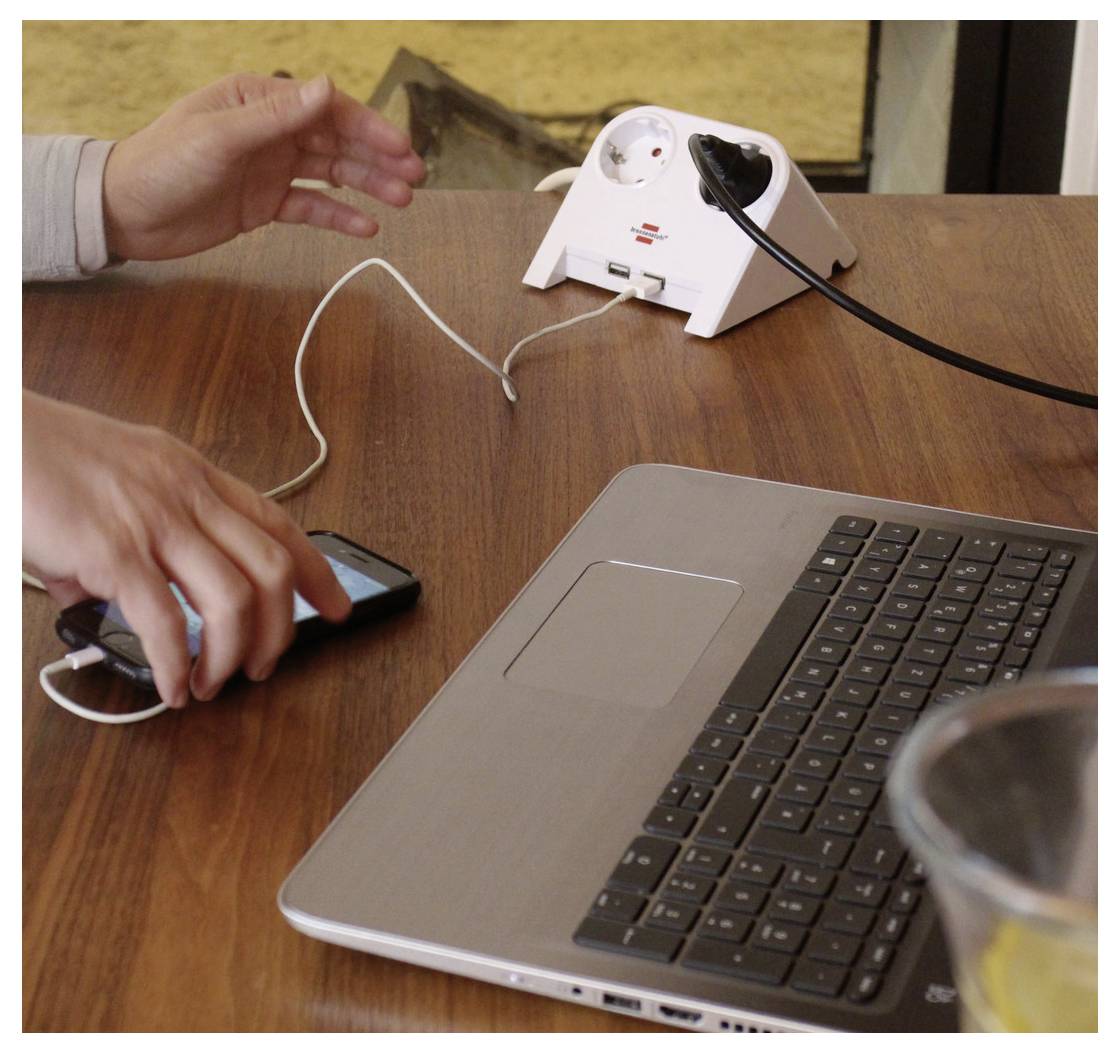 Hände laden ein Smartphone auf einem Schreibtisch neben einem Laptop auf, wobei eine weiße Steckerleiste mit mehreren eingesteckten Kabeln verwendet wird.