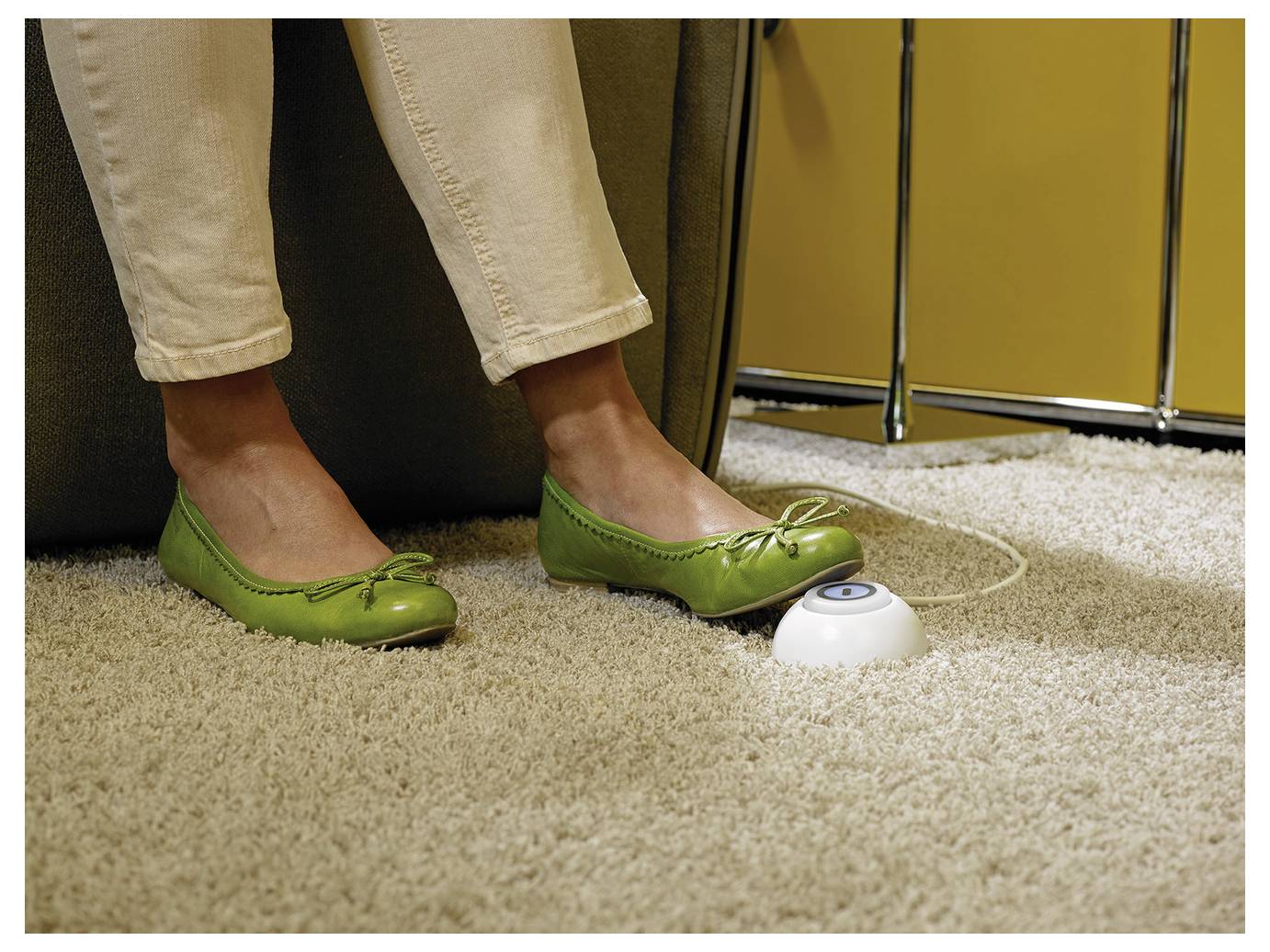 Die Füße einer Frau in grünen Ballerinas ruhen auf einem beigefarbenen Teppich neben einem kleinen weißen, knopfähnlichen Gerät, wobei ein dünnes Kabel in der Nähe liegt.