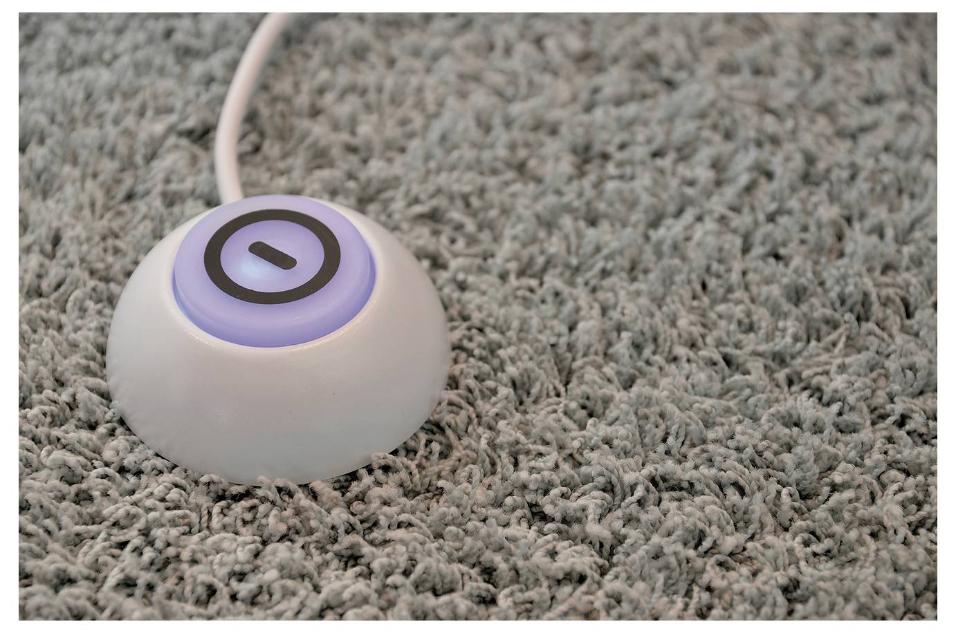 Ein kleines rundes weißes drahtloses Gerät mit einem leuchtenden violetten Ring ruht auf einem grauen Teppich.