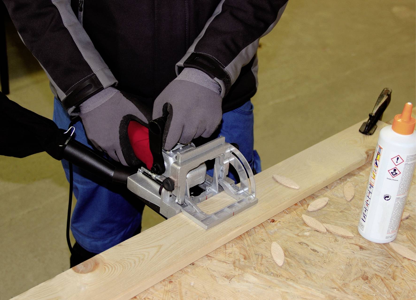 Person benutzt eine Holzbearbeitungsmaschine auf einem Brett. Daneben liegen Holzdübel und eine Klebeflasche.