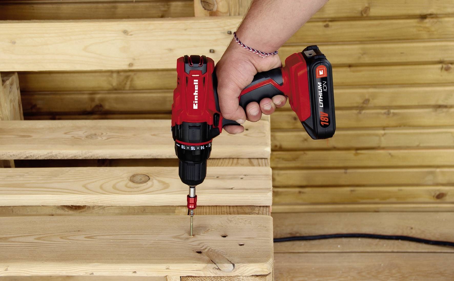 Eine Person befestigt eine Schraube in einem Holzbrett mit einem roten Akkuschrauber, im Hintergrund eine Holzverkleidung.