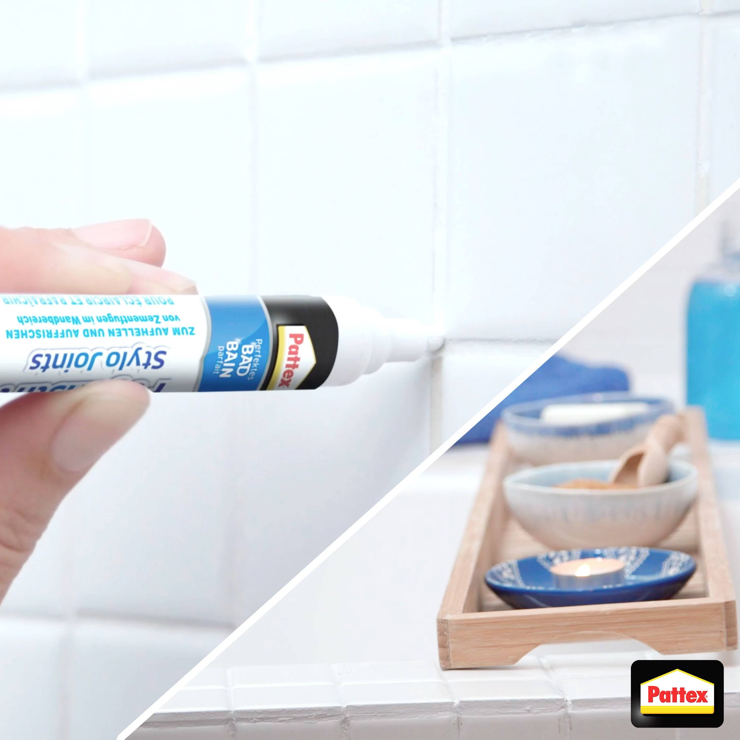 Eine Hand trägt weißen Fugenweiß-Stift auf die Fuge zwischen weißen Fliesen auf. Im Hintergrund sind blaue Accessoires sichtbar.