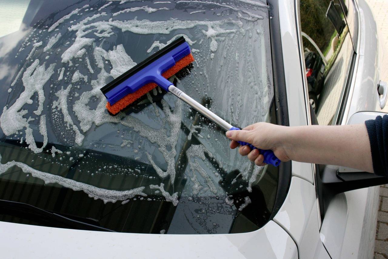 Eine Person reinigt die Windschutzscheibe eines Autos mit einem blauen Fensterabzieher. Seifenwasser ist auf dem Glas sichtbar.