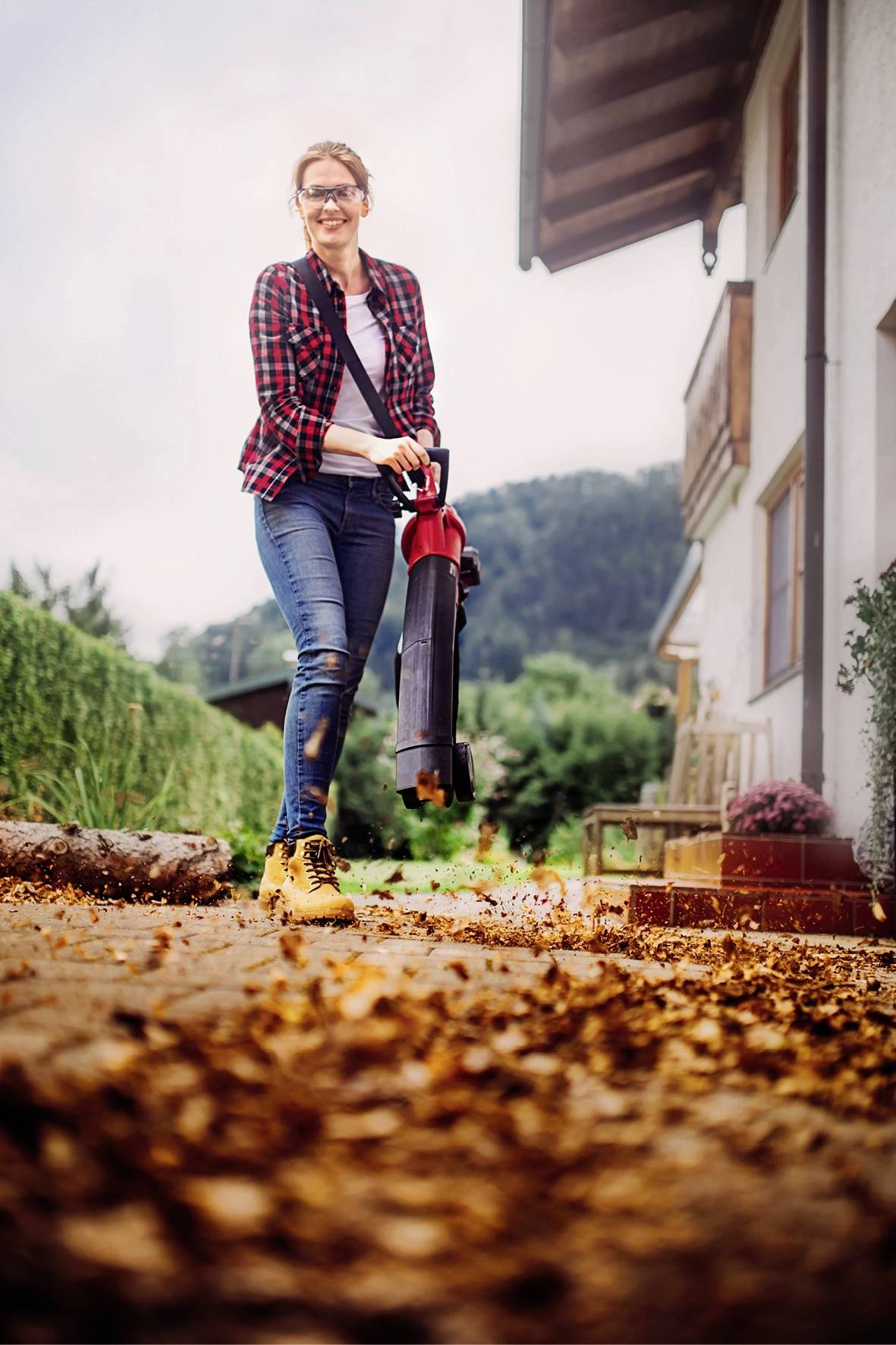 Eine Person säubert mit einem Laubbläser einen Weg neben einem Haus im Herbst. Die Umgebung ist ländlich mit grüner Vegetation.