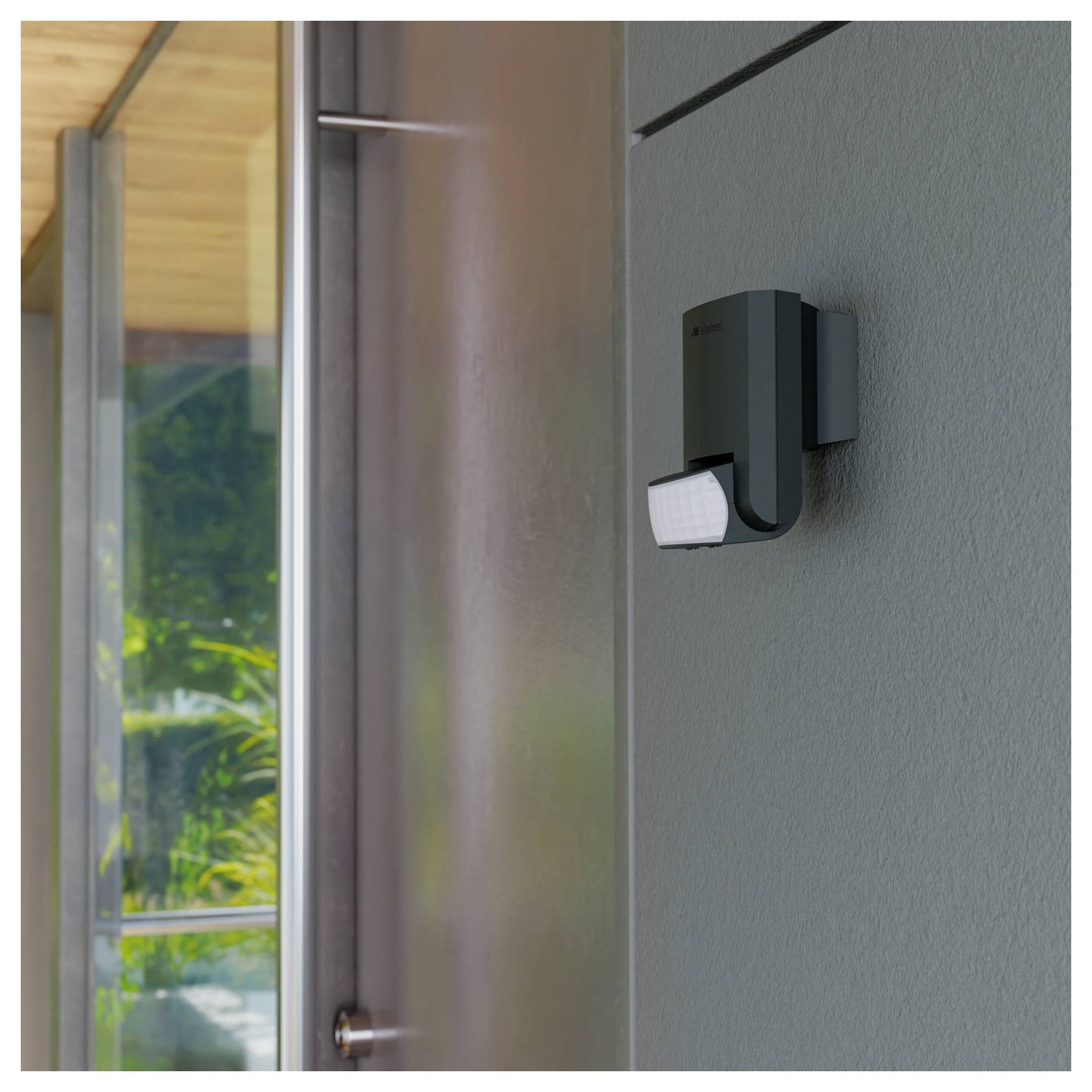 Schwarze Außenbewegungsmelder-Leuchte an einer grauen Wand neben einer Tür montiert, mit einem sichtbaren Fenster und Grünflächen in der Nähe.