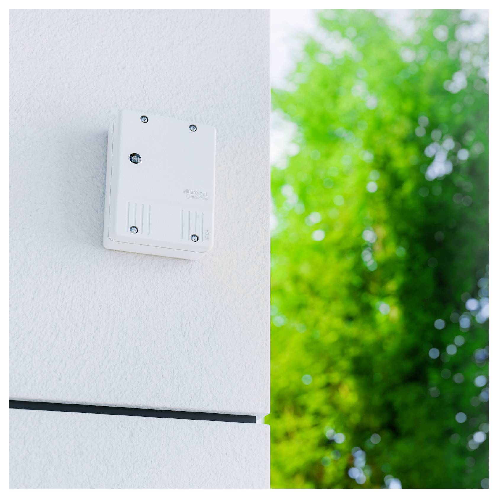 Kleiner weißer an der Wand montierter Sensor oder Steuerkasten an einer Außenwand, mit unscharfem grünem Baumwerk im Hintergrund.
