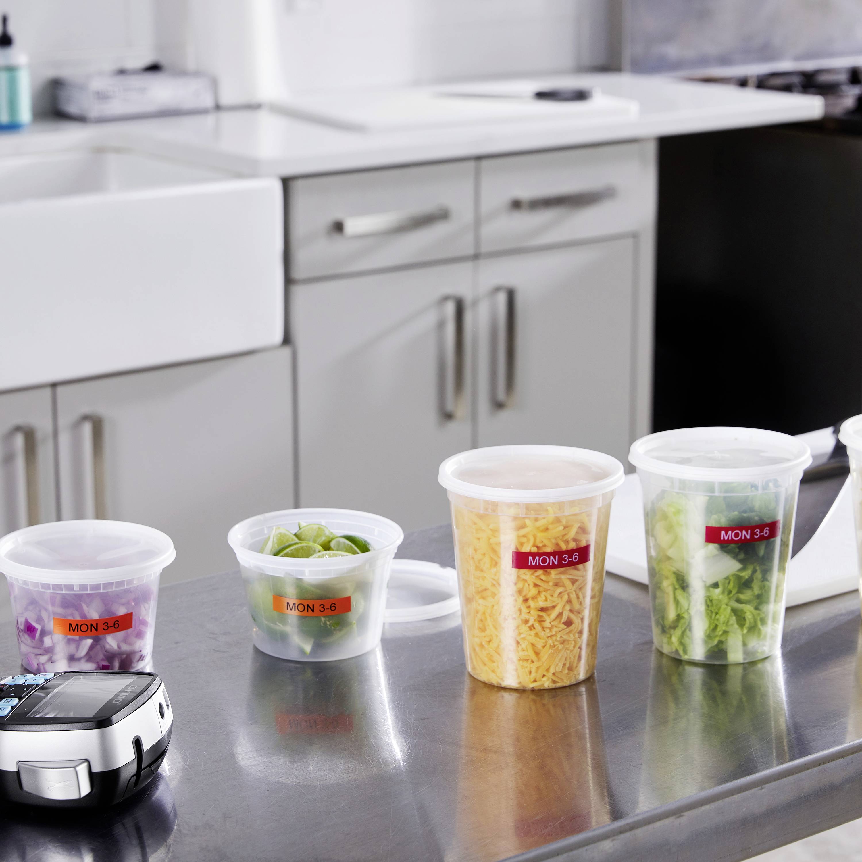 Auf einer Küchenarbeitsplatte stehen vier Plastikbehälter mit Essen, die mit 'MON 3-6' beschriftet sind. Im Vordergrund ein Gerät.
