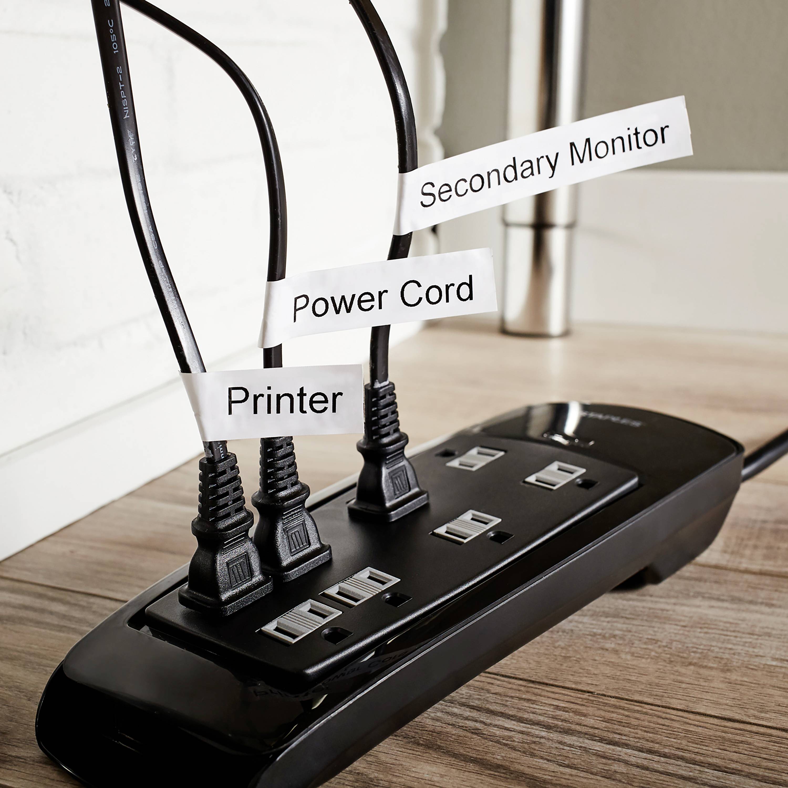 Steckdosenleiste auf Holzboden mit drei eingesteckten Kabeln; beschriftet mit 'Secondary Monitor', 'Power Cord', 'Printer'.
