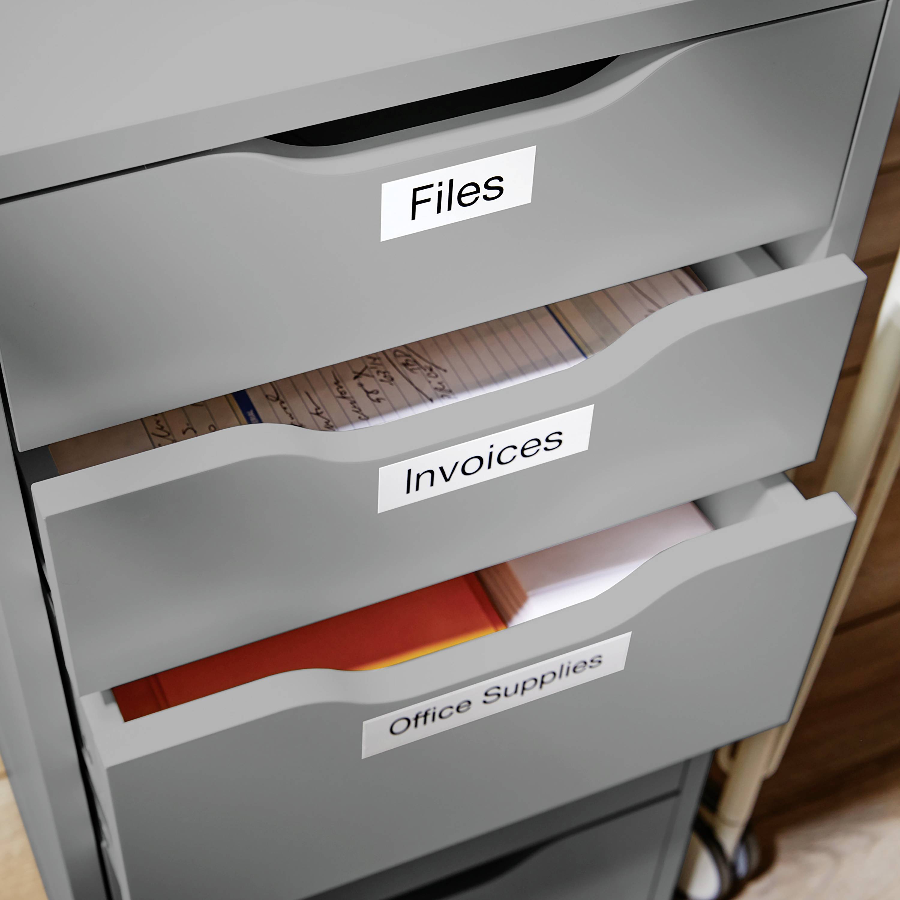 Ein grauer Aktenschrank mit drei geöffneten Schubladen, beschriftet mit 'Files', 'Invoices' und 'Office Supplies', gefüllt mit Papieren.