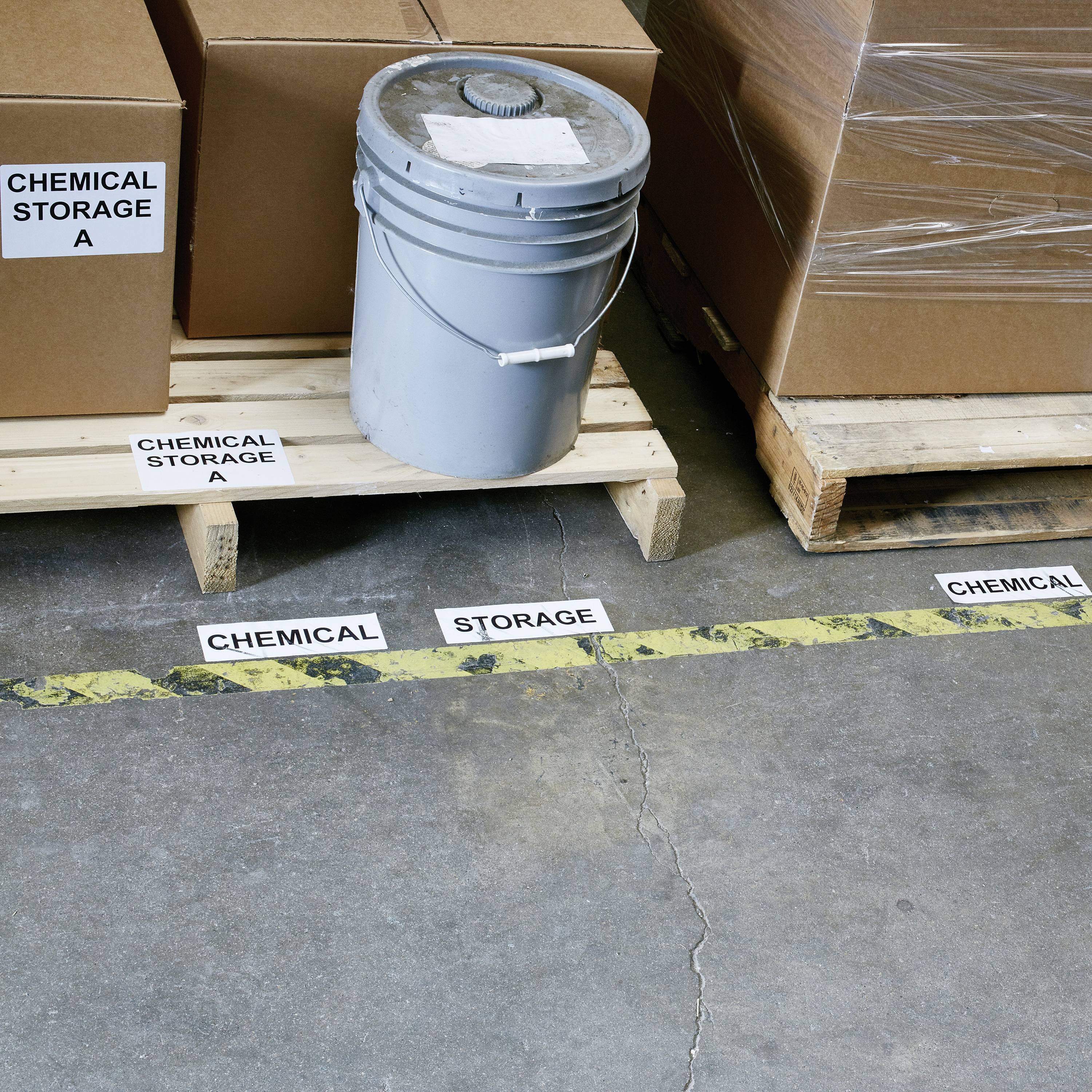 Ein Lager mit beschrifteten Bodenschildern: 'CHEMICAL STORAGE', Kartons und ein Plastikeimer auf einer Palette.