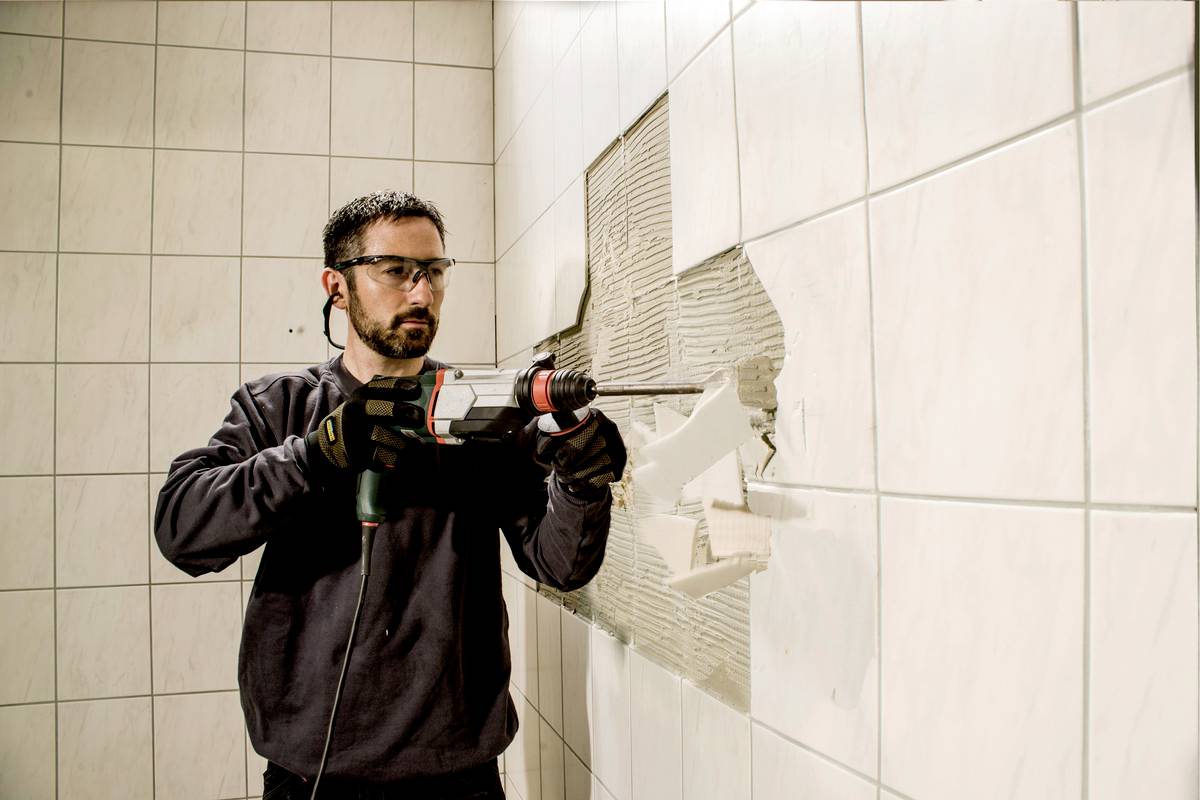 Arbeiter mit Schutzbrille und Handschuhen bohrt mit einer Maschine Fliesen von einer Wand ab.