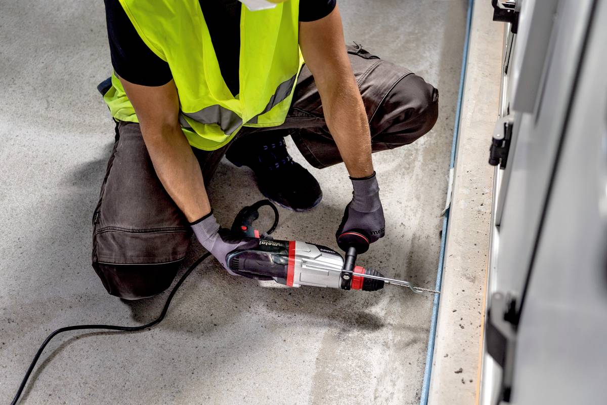 Person in gelber Weste und Handschuhen benutzt eine Bohrmaschine, um ein Loch in den Boden neben einer Wand zu bohren.