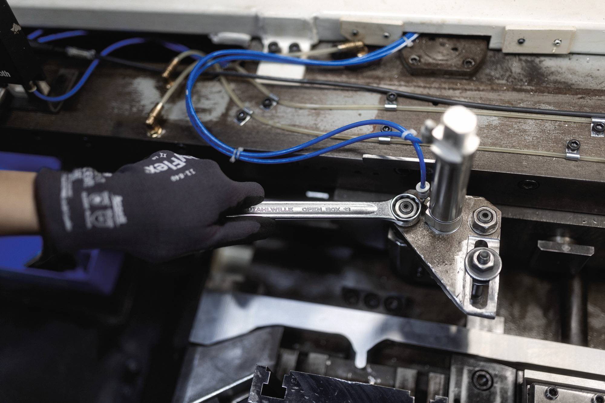 Eine behandschuhte Hand stellt mit einem Schraubenschlüssel ein mechanisches Bauteil an einer Industriemaschine ein, die mit blauen Drähten verbunden ist, in einer Fabrikumgebung.