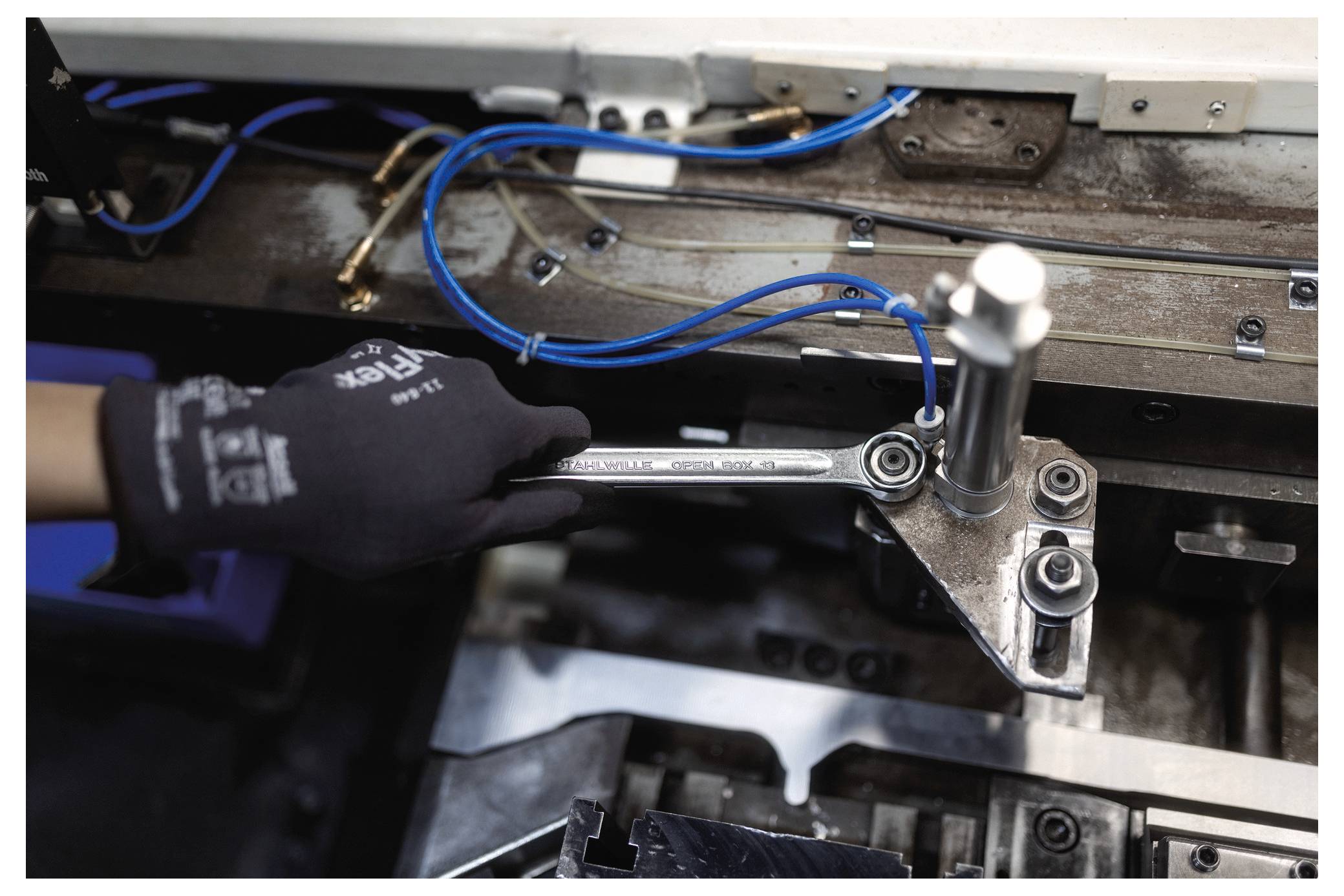 Die behandschuhte Hand einer Person bedient einen Schraubenschlüssel an einer Industriemaschine mit sichtbaren blauen Kabeln, was auf Wartungs- oder Reparaturarbeiten hindeutet.