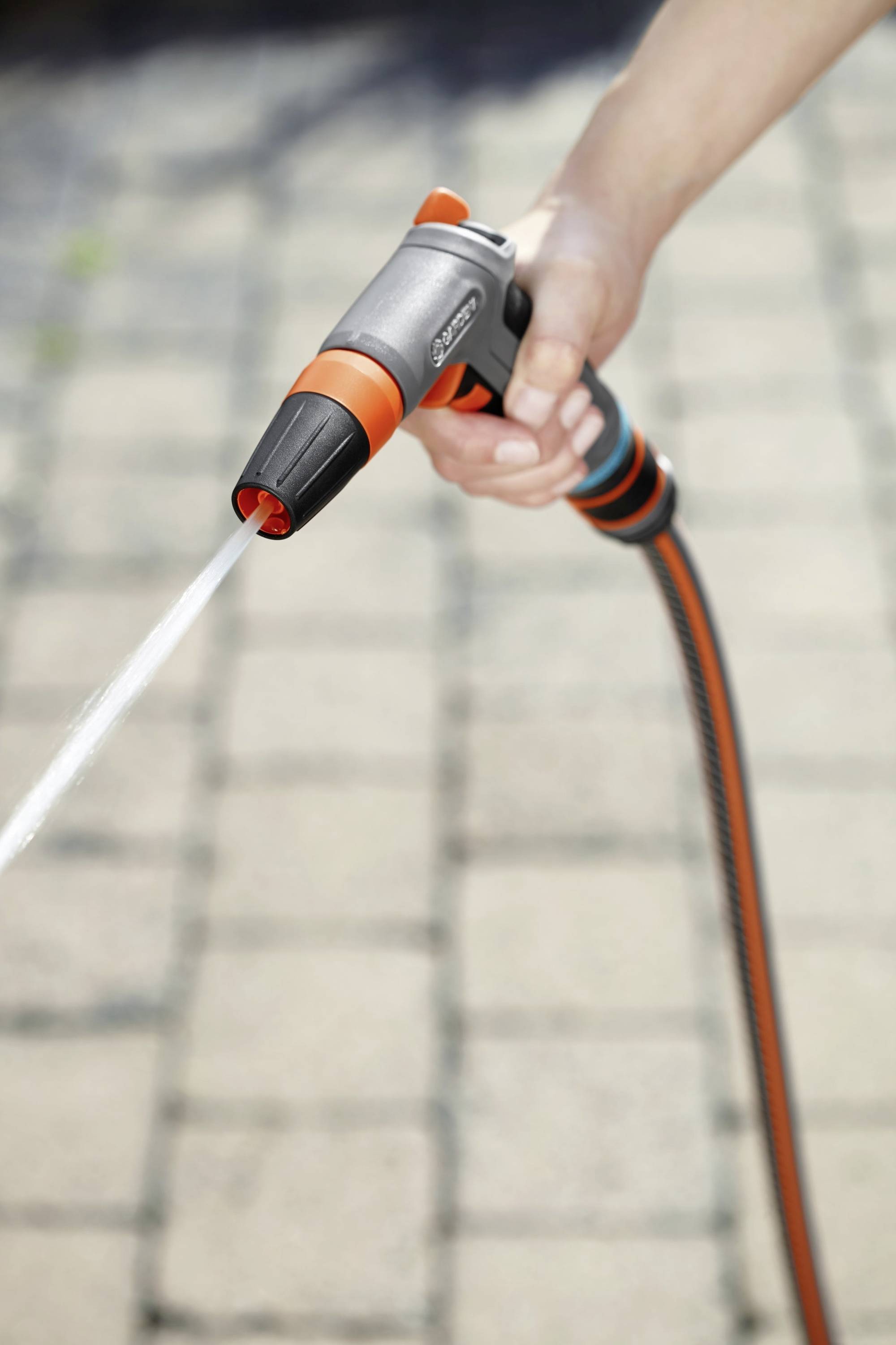 Eine Hand hält einen Gartenschlauch mit einer Sprühdüse, Wasserstrahl spritzt auf einen gepflasterten Boden.