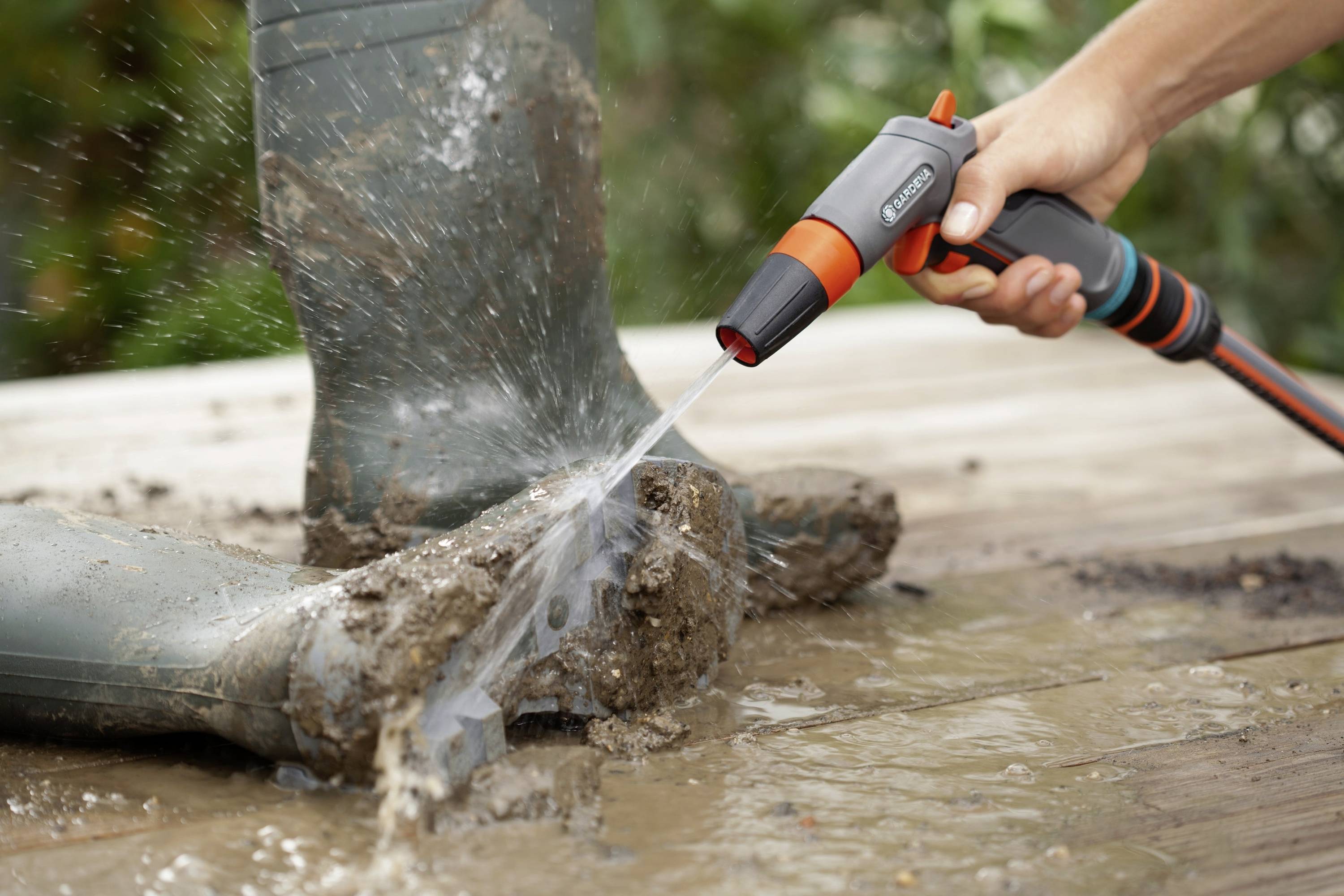 Eine Person spritzt mit einem Gartenschlauch Wasser auf stark verschmutzte Gummistiefel, um den anhaftenden Schlamm zu entfernen.