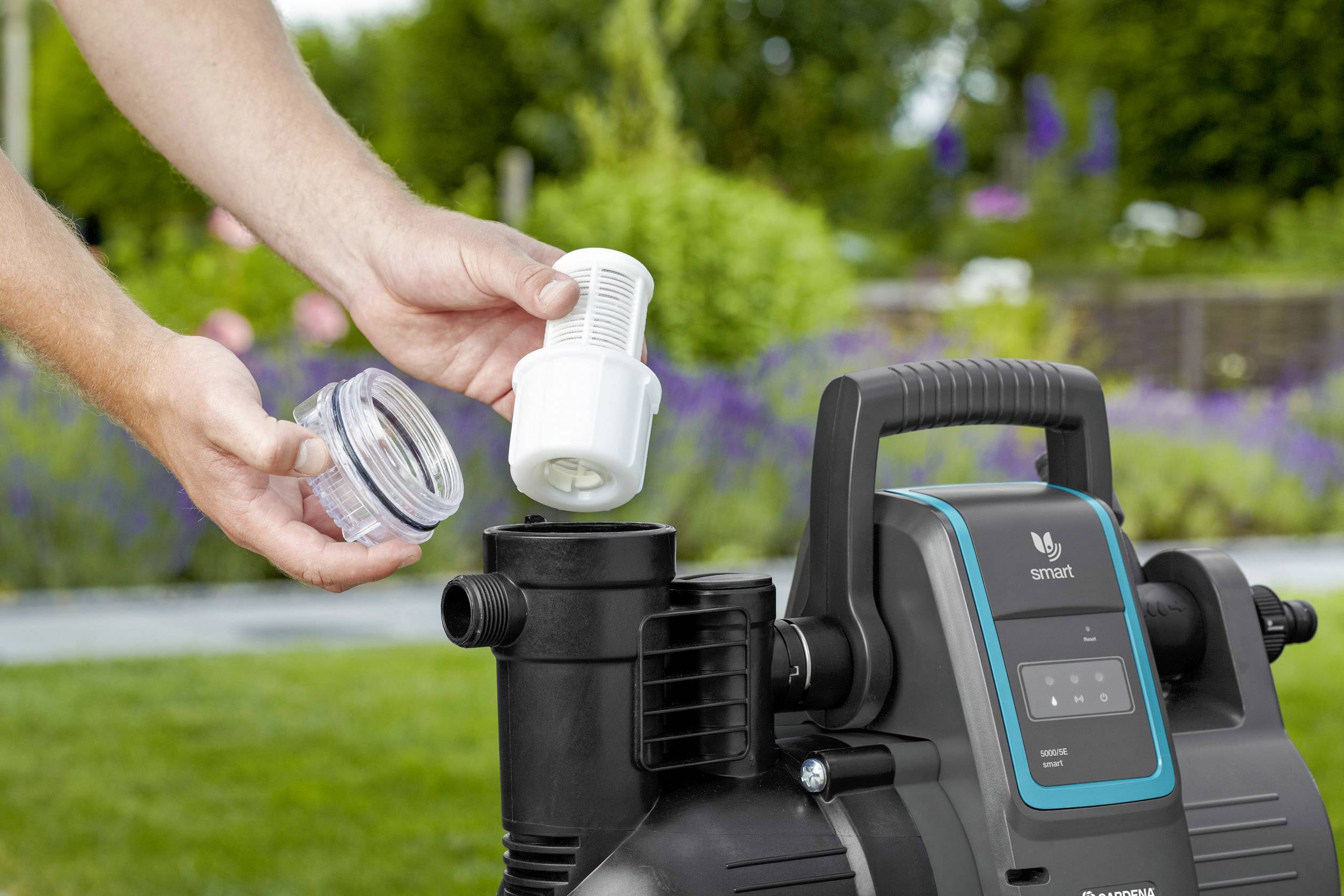 Eine Person reinigt den Filter einer schwarzen Wasserpumpe im Garten. Die Pumpe hat ein digitales Display und befindet sich im Freien.