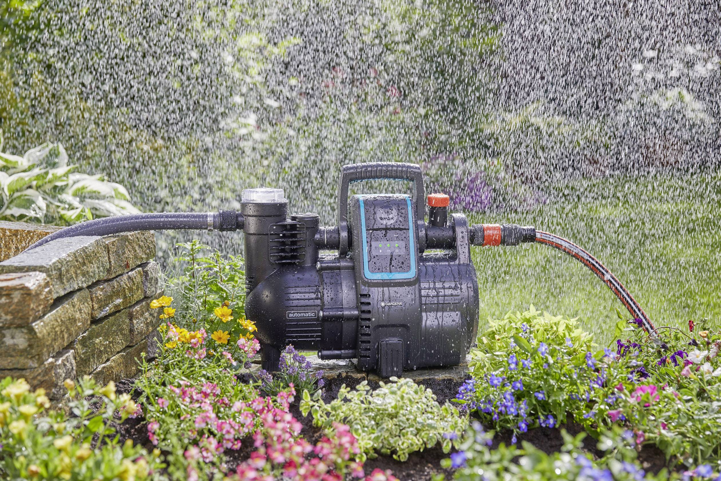 Eine Gartenpumpe bewässert ein Blumenbeet in einem grünen Garten. Blumen in verschiedenen Farben blühen um die Pumpe herum.