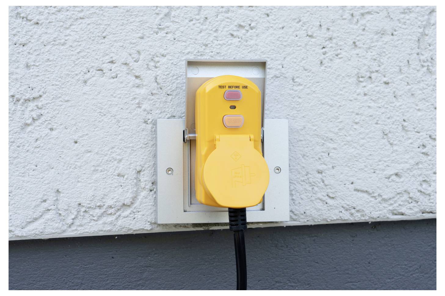 Gelbes Schutzdach für Außensteckdosen an einer Wand montiert, mit einem Stromkabel, das eingesteckt ist und nach unten hängt.