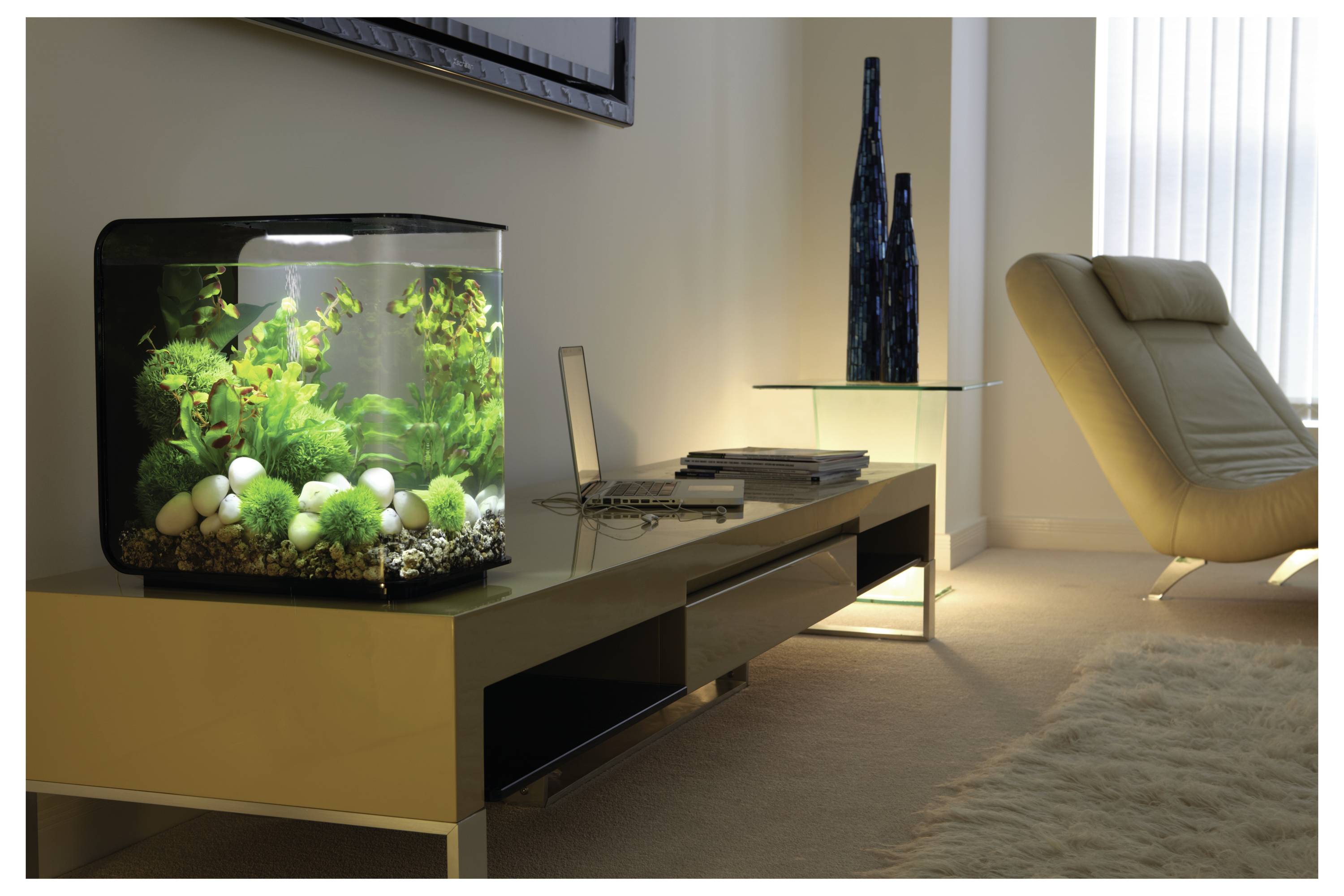 Ein moderner Wohnraum präsentiert einen eleganten beigen Liegestuhl, einen niedrigen Tisch mit einem Laptop und ein beleuchtetes Aquarium mit leuchtend grünen Pflanzen.
