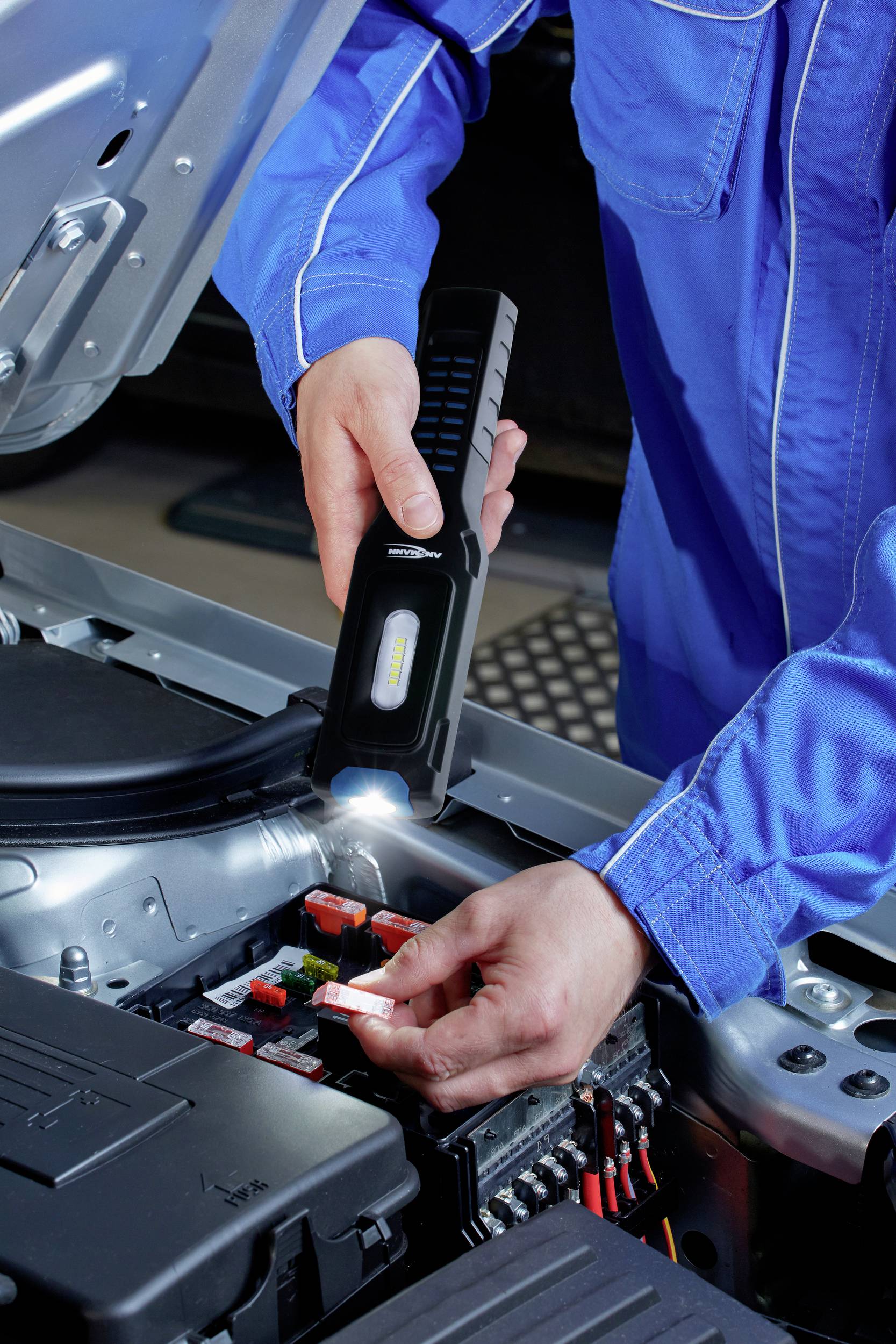 Person in blauer Arbeitskleidung überprüft die Sicherungen in einem offenen Automotor mit einer Taschenlampe.