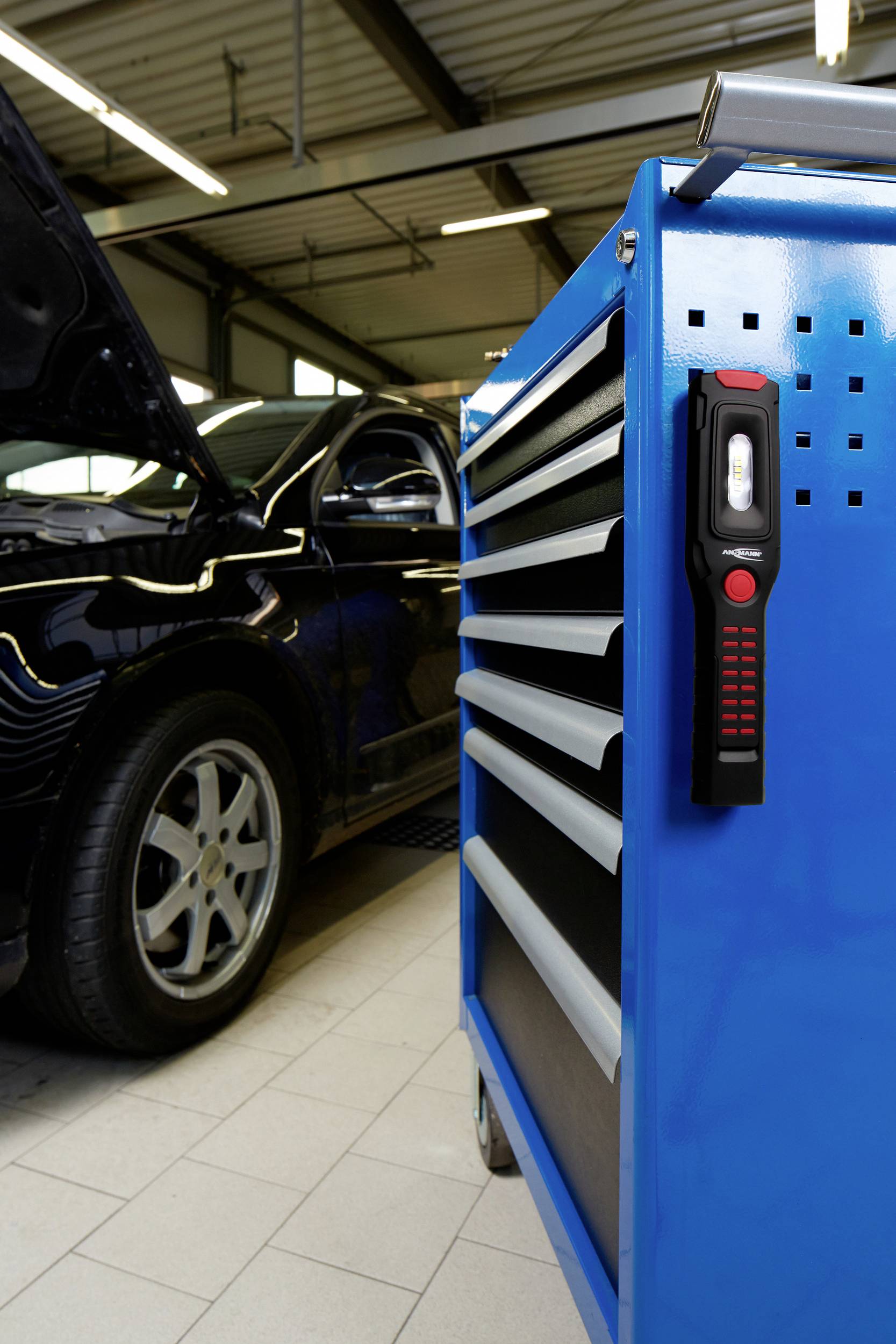 Ein schwarzes Auto mit geöffneter Motorhaube steht in einer Werkstatt. Im Vordergrund ist ein blauer Werkzeugwagen mit Lampe zu sehen.