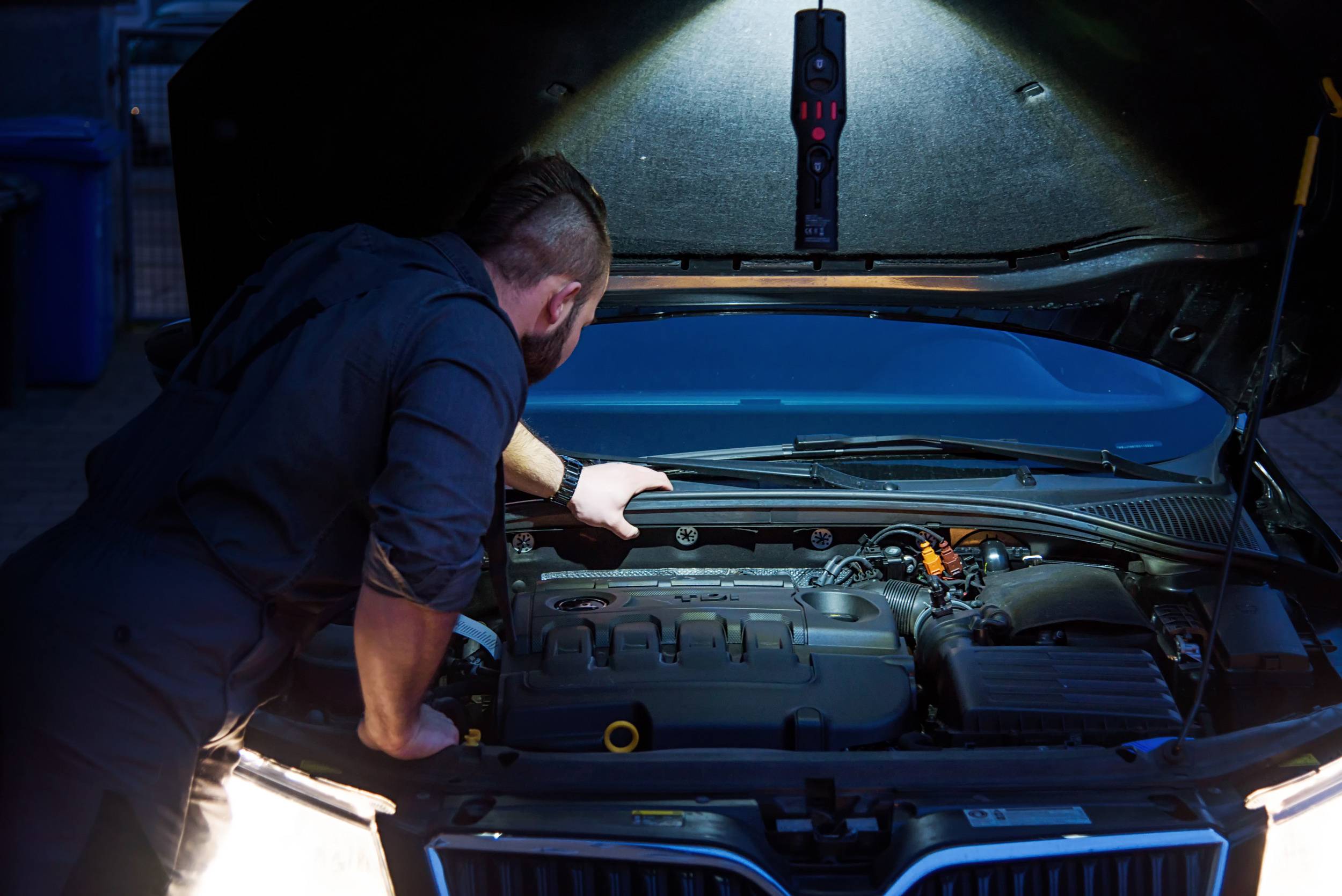 Ein Mechaniker inspiziert den Motorraum eines Autos unter einer geöffneten Motorhaube. Eine Lampe beleuchtet den Bereich.