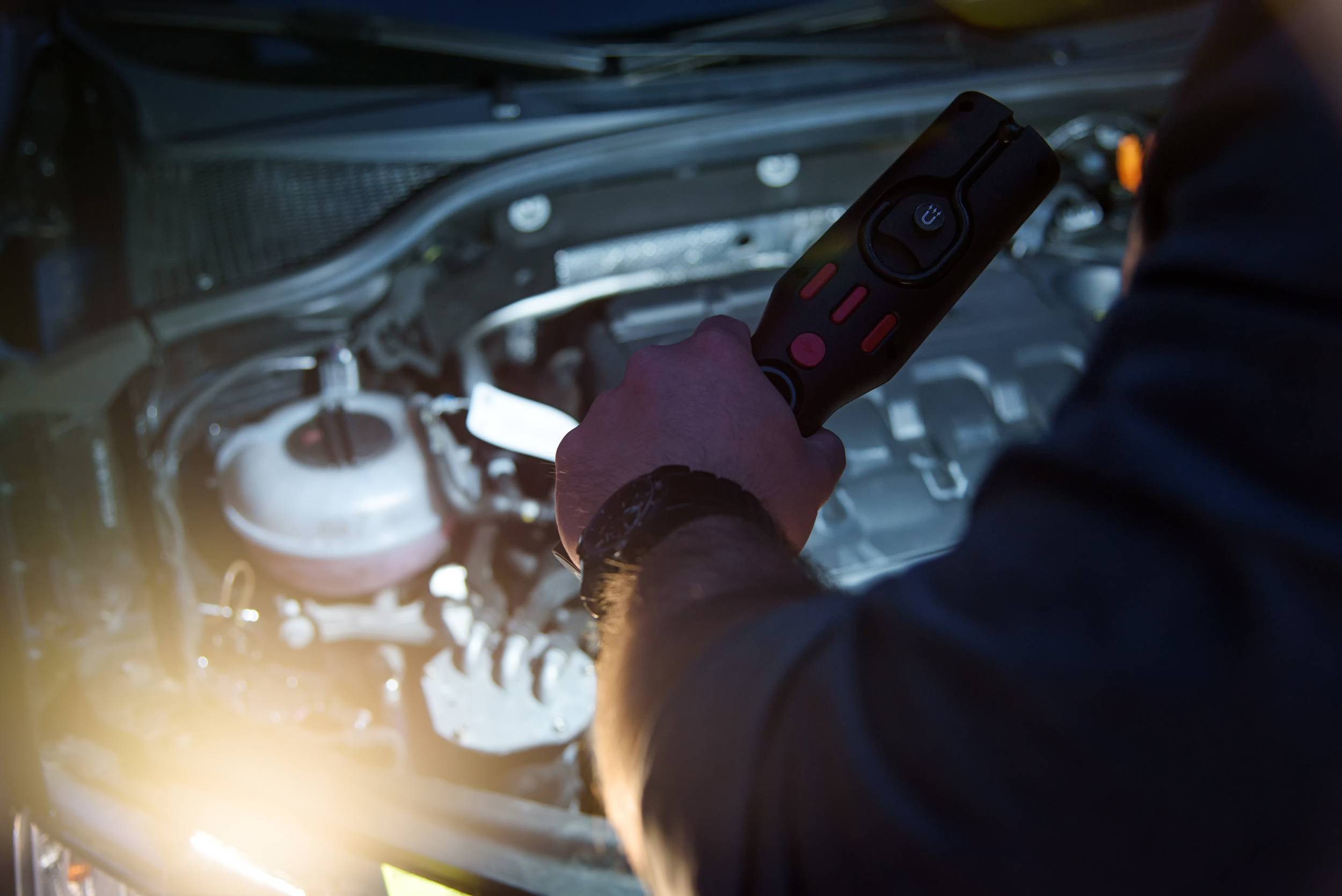 Eine Person hält ein Diagnosegerät über einen geöffneten Auto-Motorraum, um technische Probleme zu analysieren oder zu reparieren.