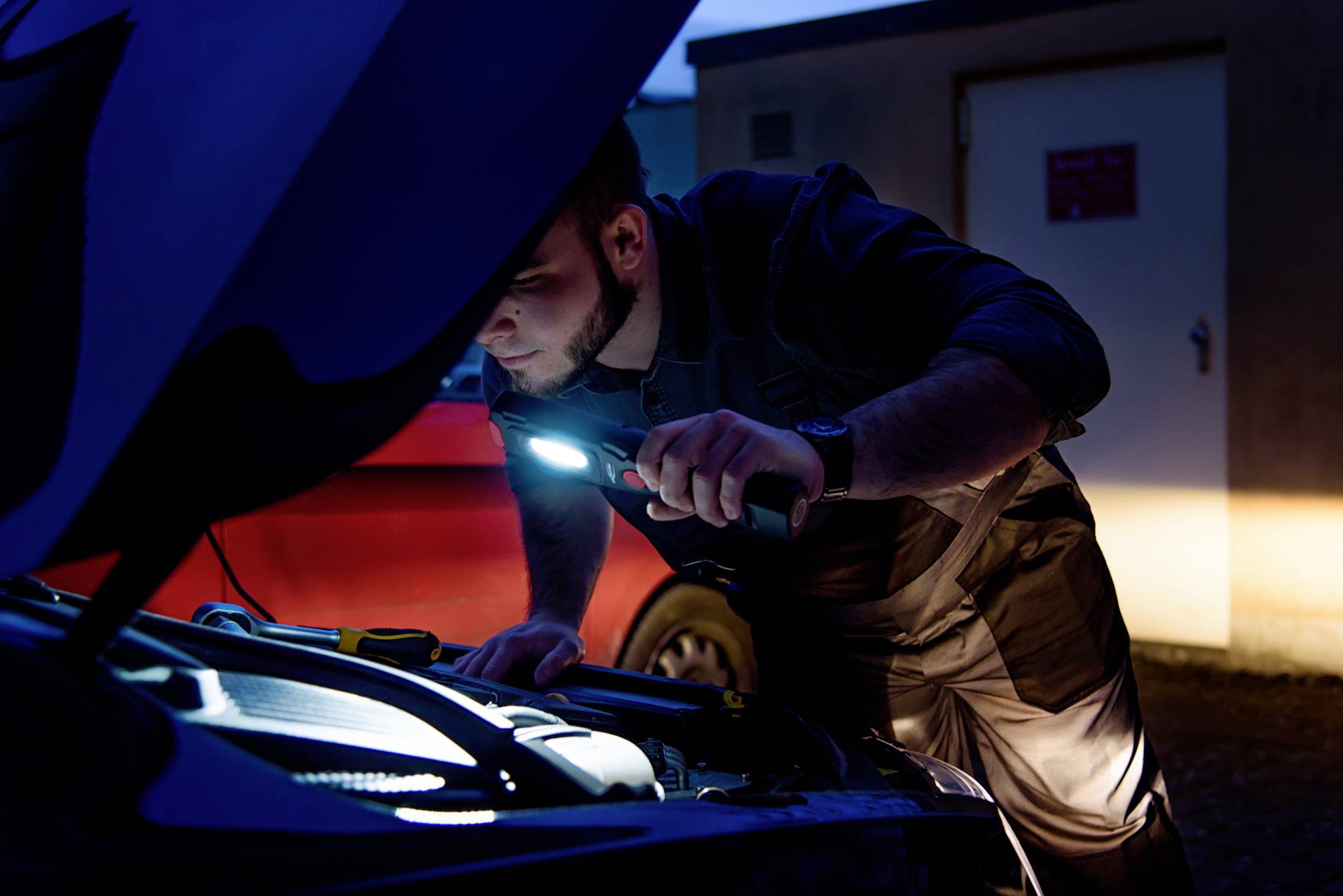 Eine Person untersucht mit einer Taschenlampe den Motorraum eines Autos bei Dämmerung.