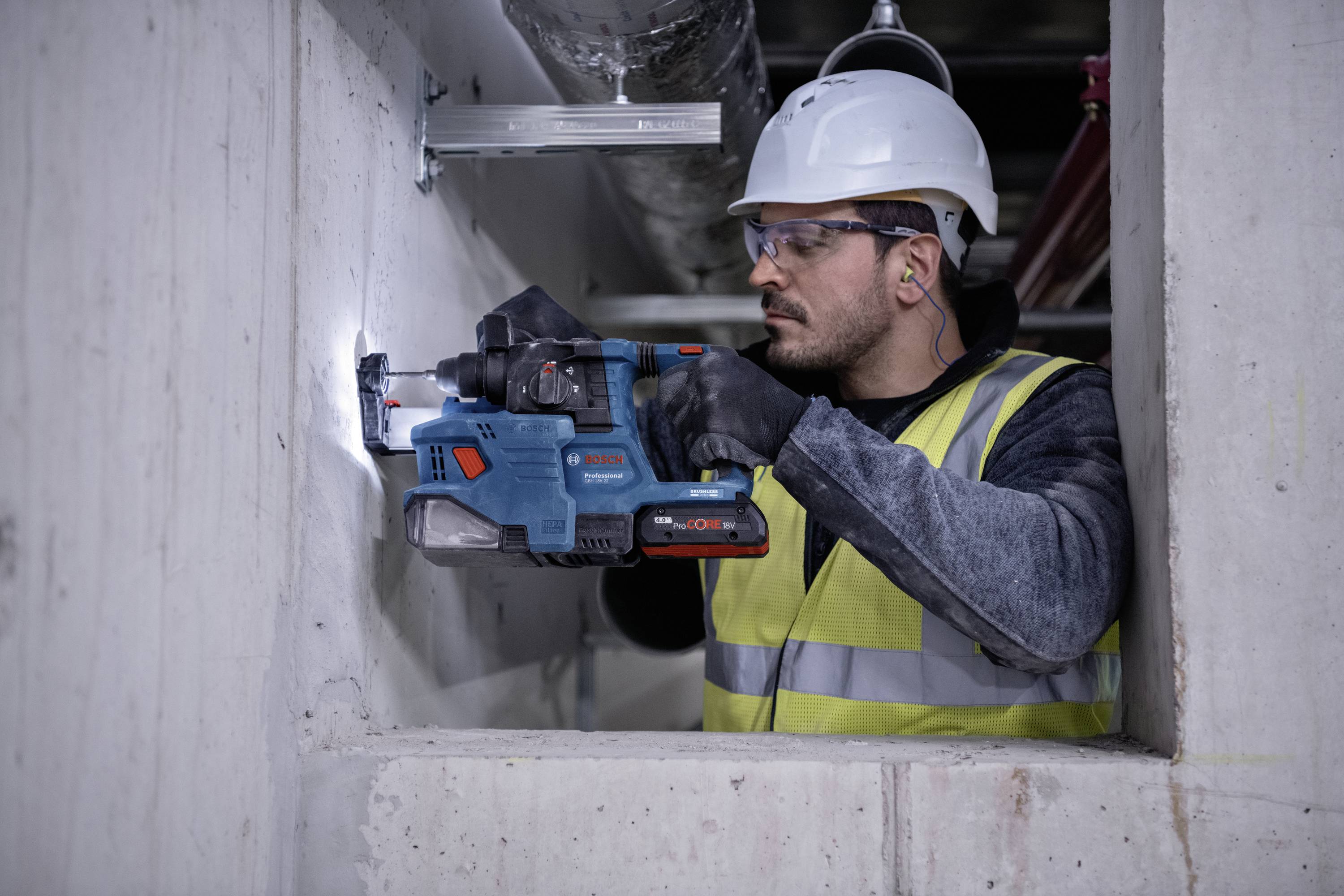 Ein Bauarbeiter trägt Helm und Schutzweste und bohrt mit einer elektrischen Bohrmaschine ein Loch in eine Betonwand.