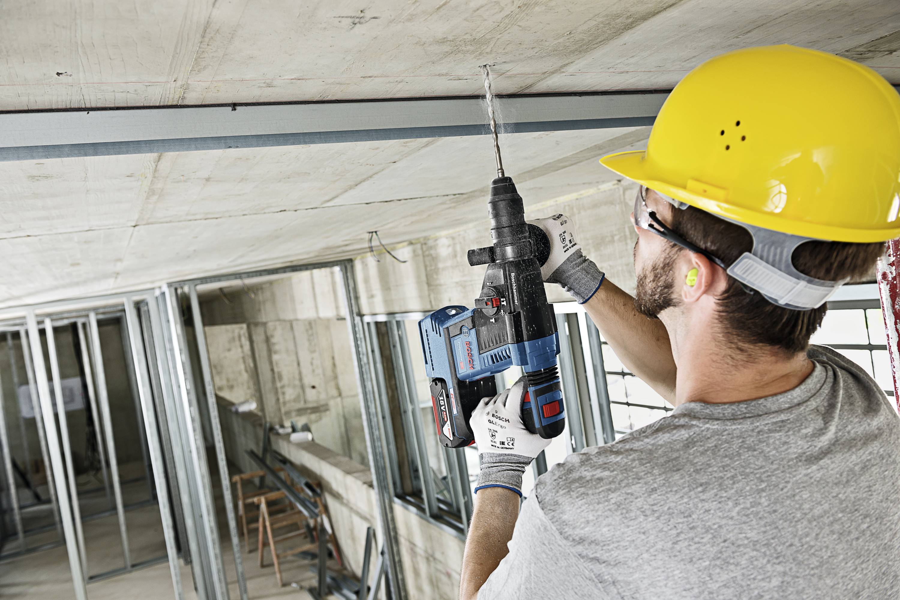 Arbeiter bohrt mit einer elektrischen Bohrmaschine in die Decke einer Baustelle. Trägt gelben Schutzhelm und Ohrstöpsel.