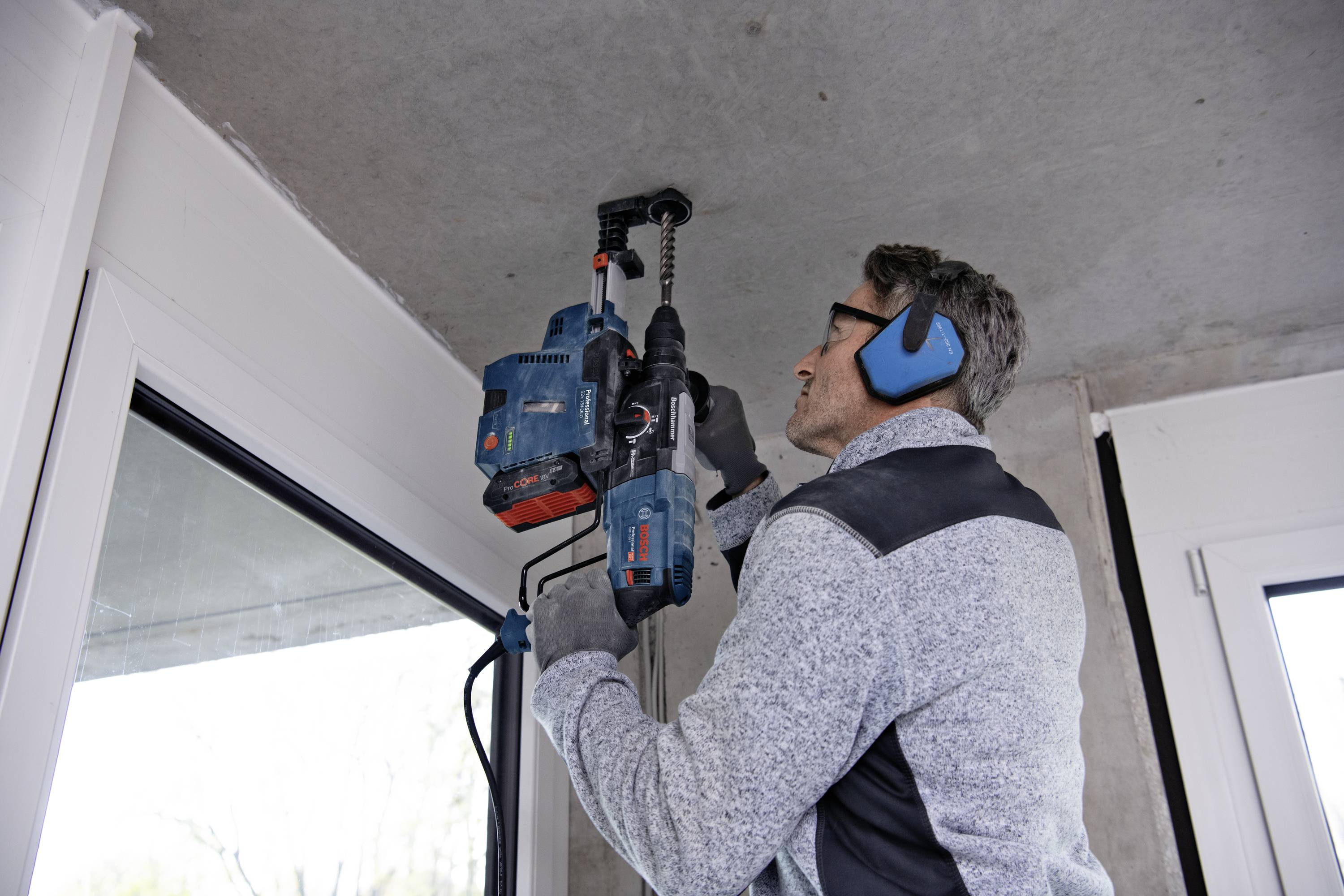 Ein Mann verwendet eine Bohrmaschine, um in die Betondecke über einem Fenster zu bohren. Er trägt Gehörschutz und Handschuhe.