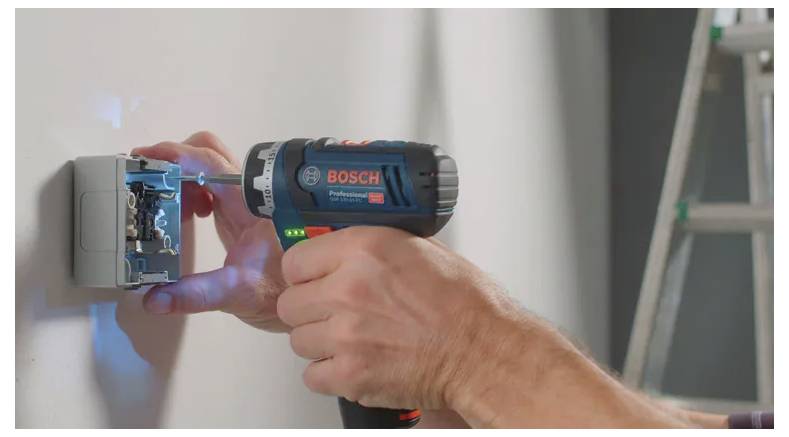 Eine Person verwendet einen Bosch-Akku-Bohrer, um an einer Stromverteilerdose zu arbeiten, die an einer Wand montiert ist. Eine Leiter ist im Hintergrund zu sehen.