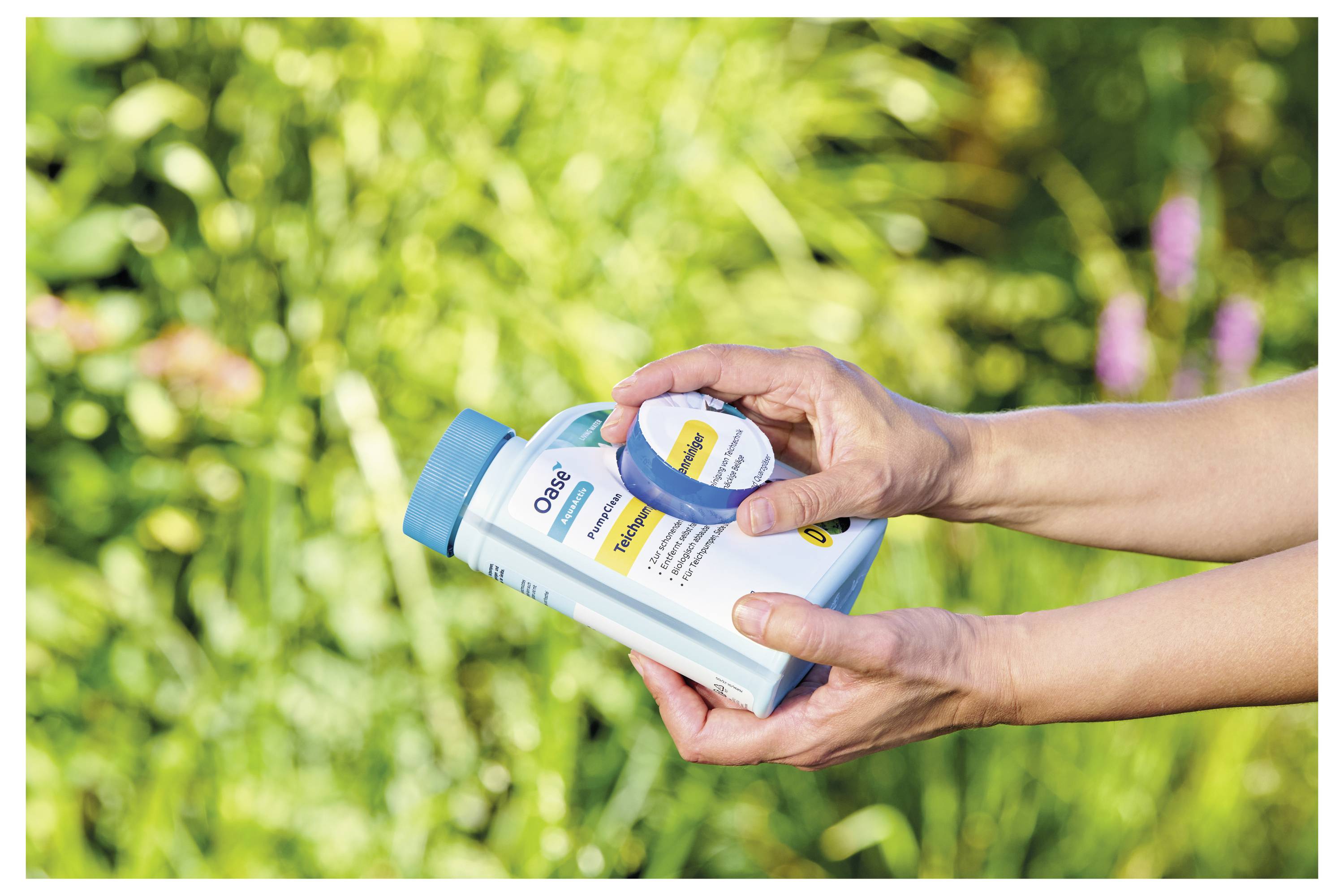 Hände, die einen Behälter mit Dünger vor einem grünen Gartenhindergrund halten.