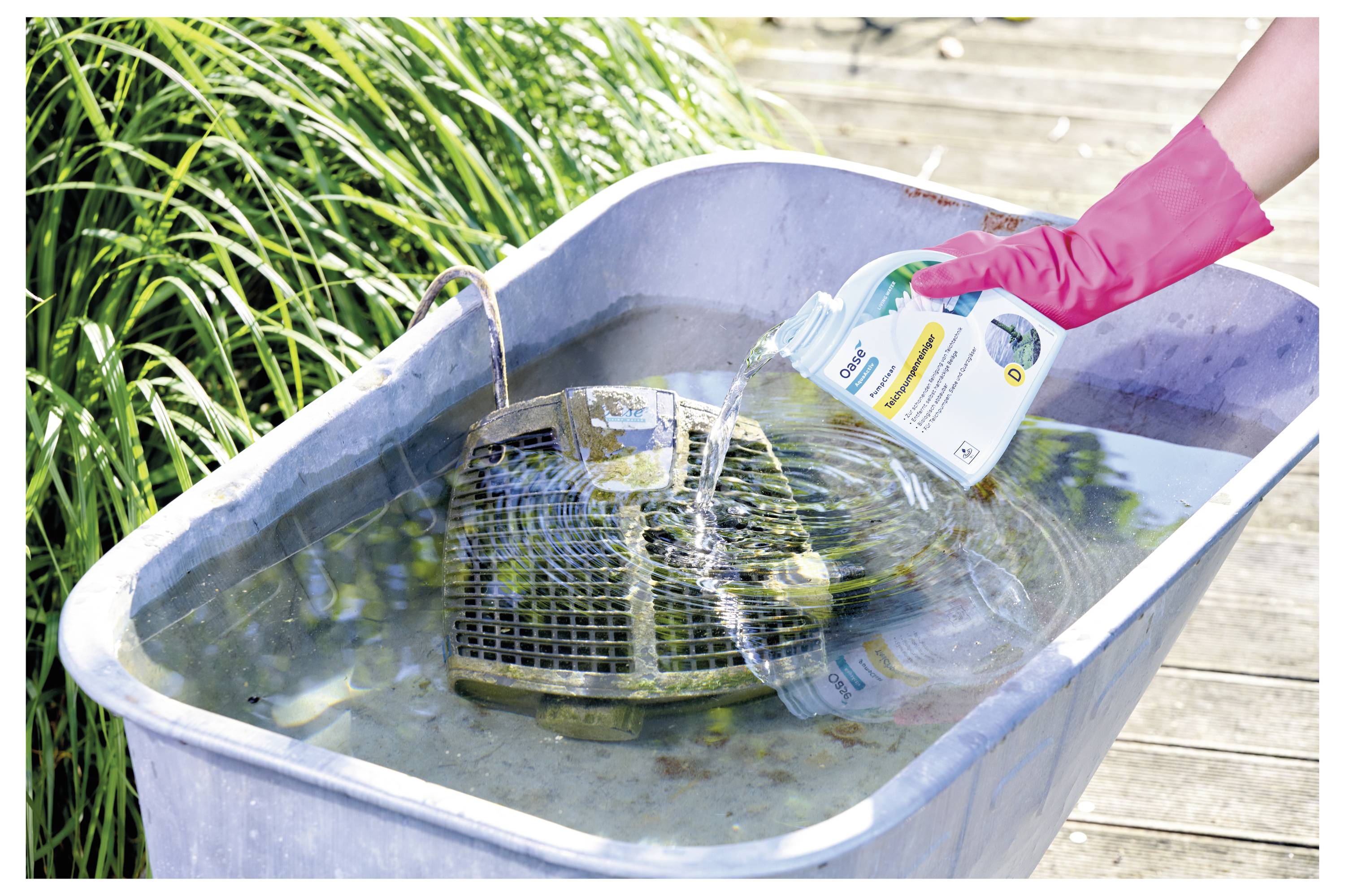 Eine Person in einem pinken Handschuh gießt Reinigungsmittel in eine Wanne, in der ein verschmutzte Grillrost in Wasser einweicht, daneben Gras und eine Holzterrasse.