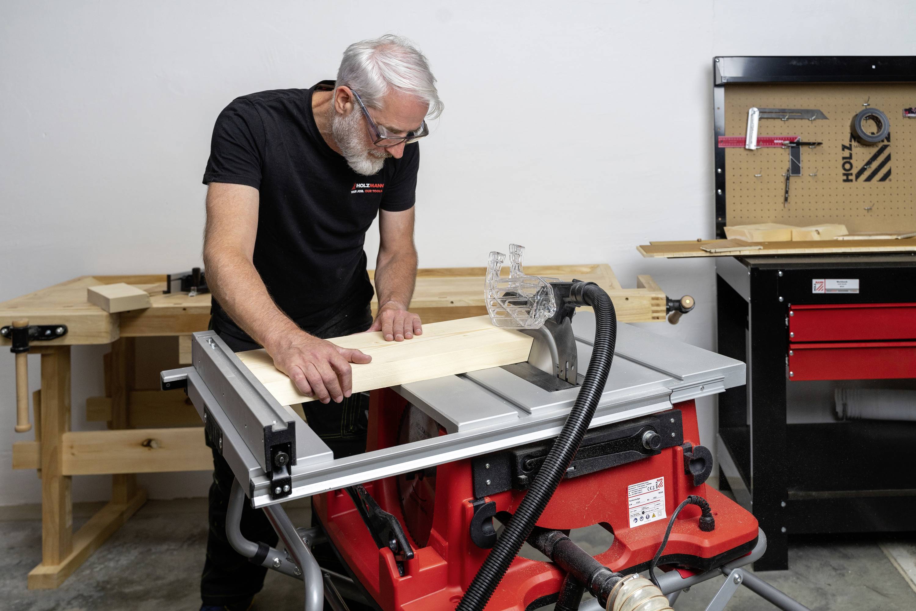 Eine Person in einer Werkstatt sägt ein Holzbrett mit einer Tischkreissäge. Im Hintergrund sind Werkzeuge an einer Wandtafel zu sehen.