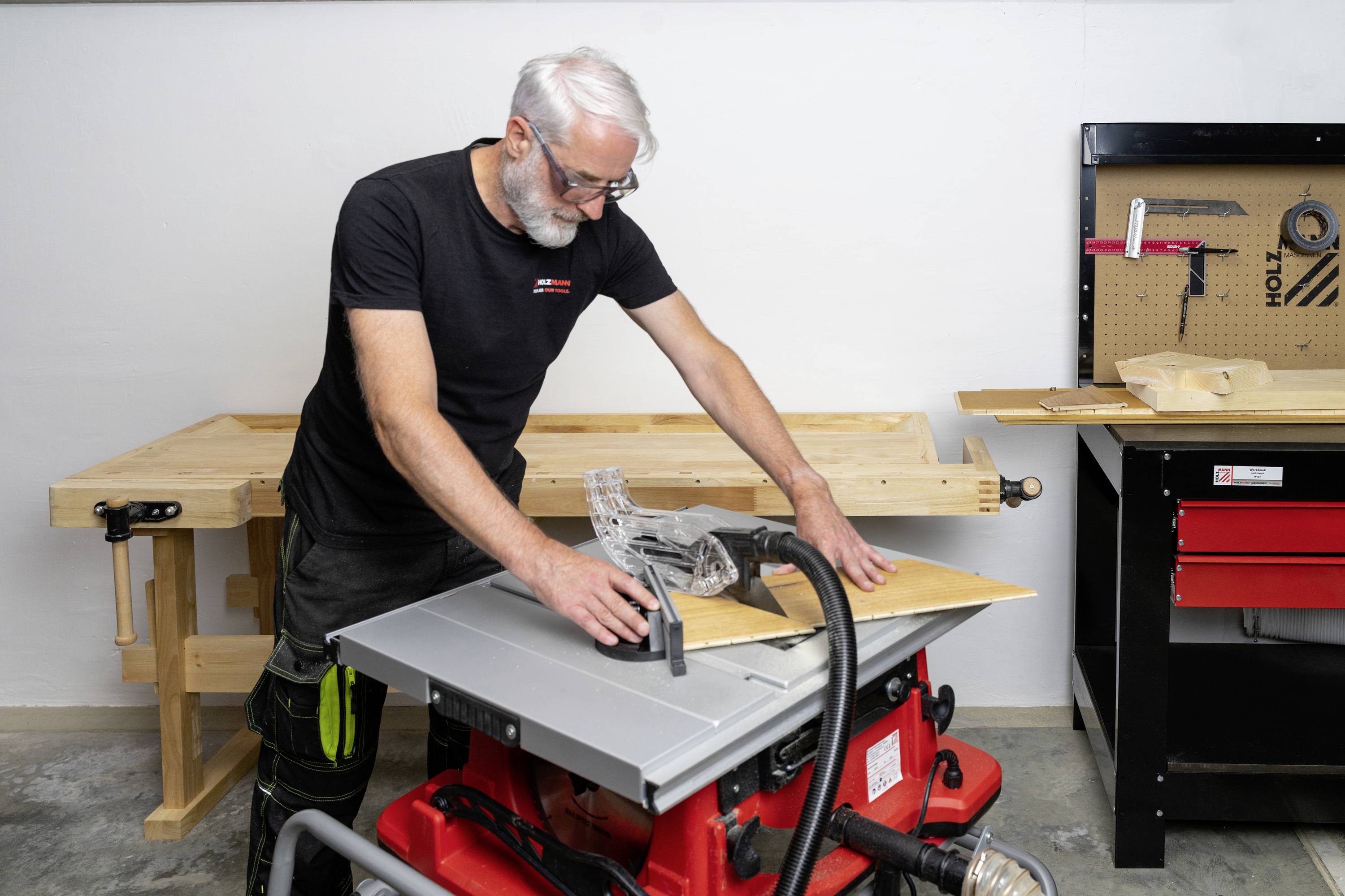 Ein Mensch mit weißem Haar arbeitet an einer Tischkreissäge in einer Werkstatt. Im Hintergrund sind Werkbänke und Werkzeuge zu sehen.