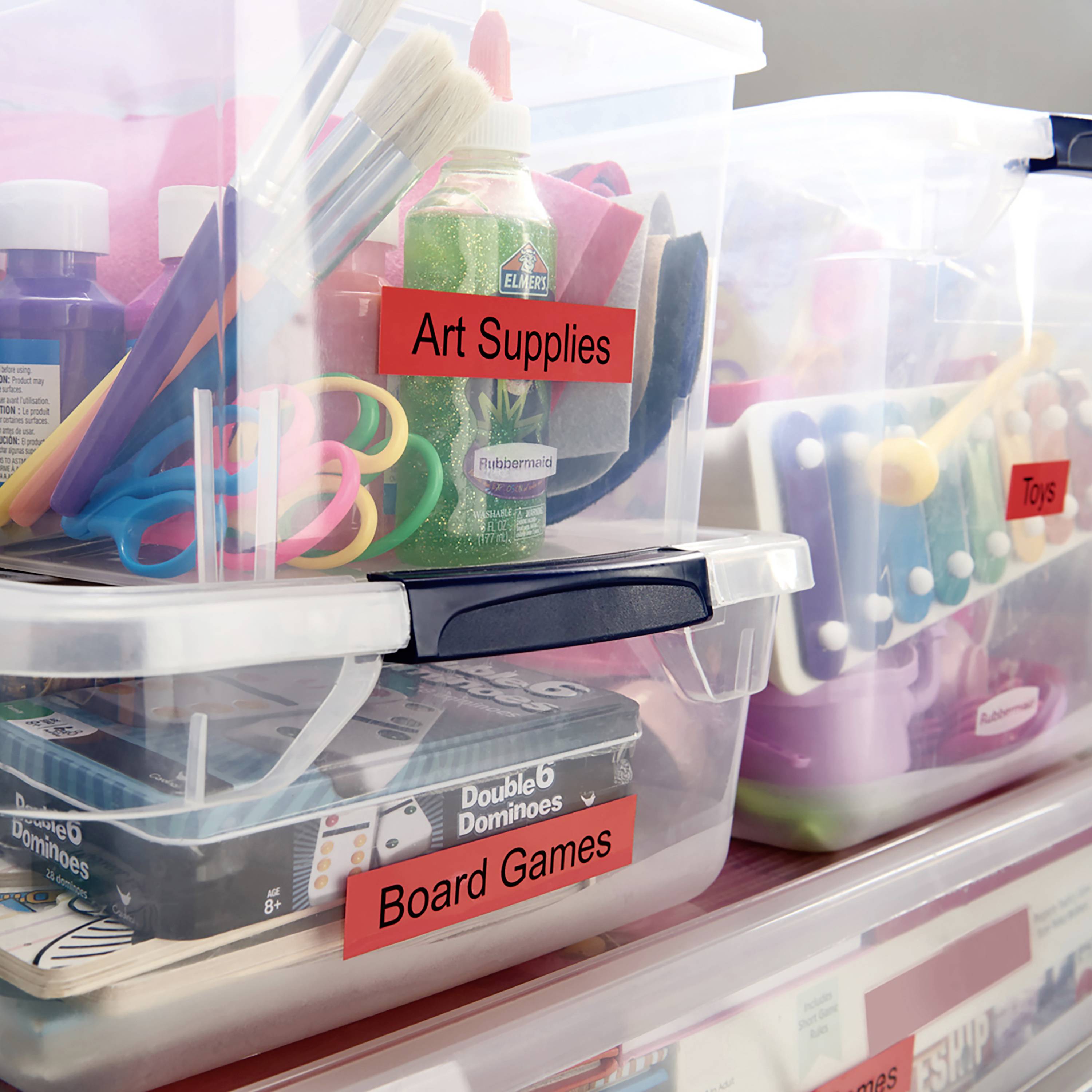 Kunstbedarf und Spielsachen in transparenten Boxen beschriftet mit 'Art Supplies', 'Toys', 'Board Games' und 'Double 9 Dominoes'.