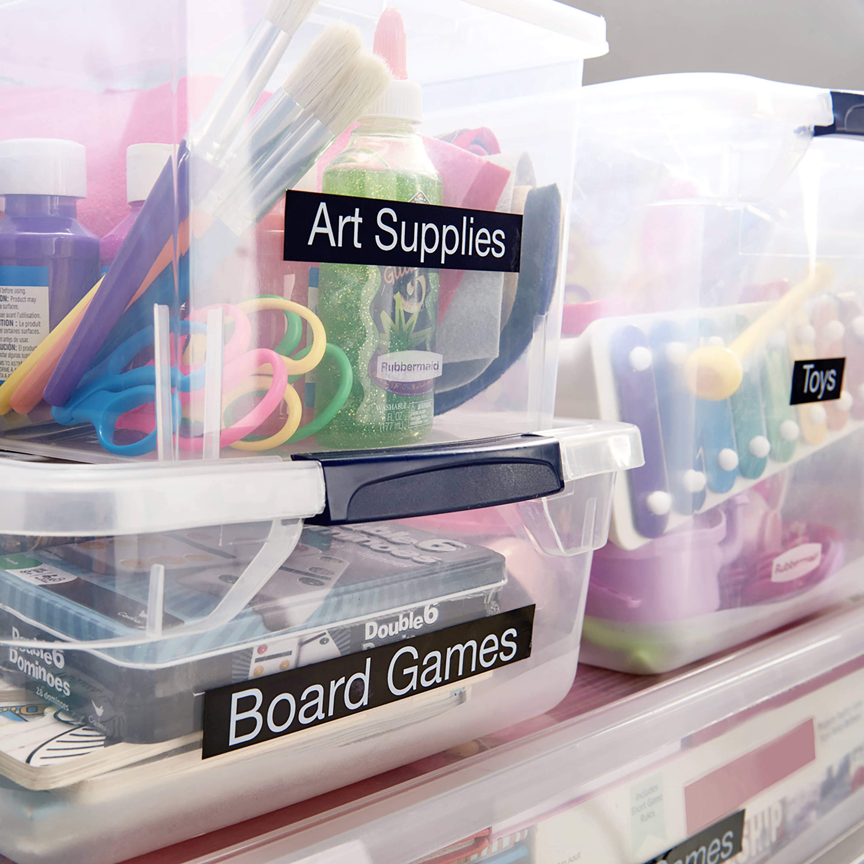 Kunstbedarf, Brettspiele und Spielzeug in beschrifteten Plastikboxen zur organisierten Lagerung. Pinsel und Farben sichtbar.