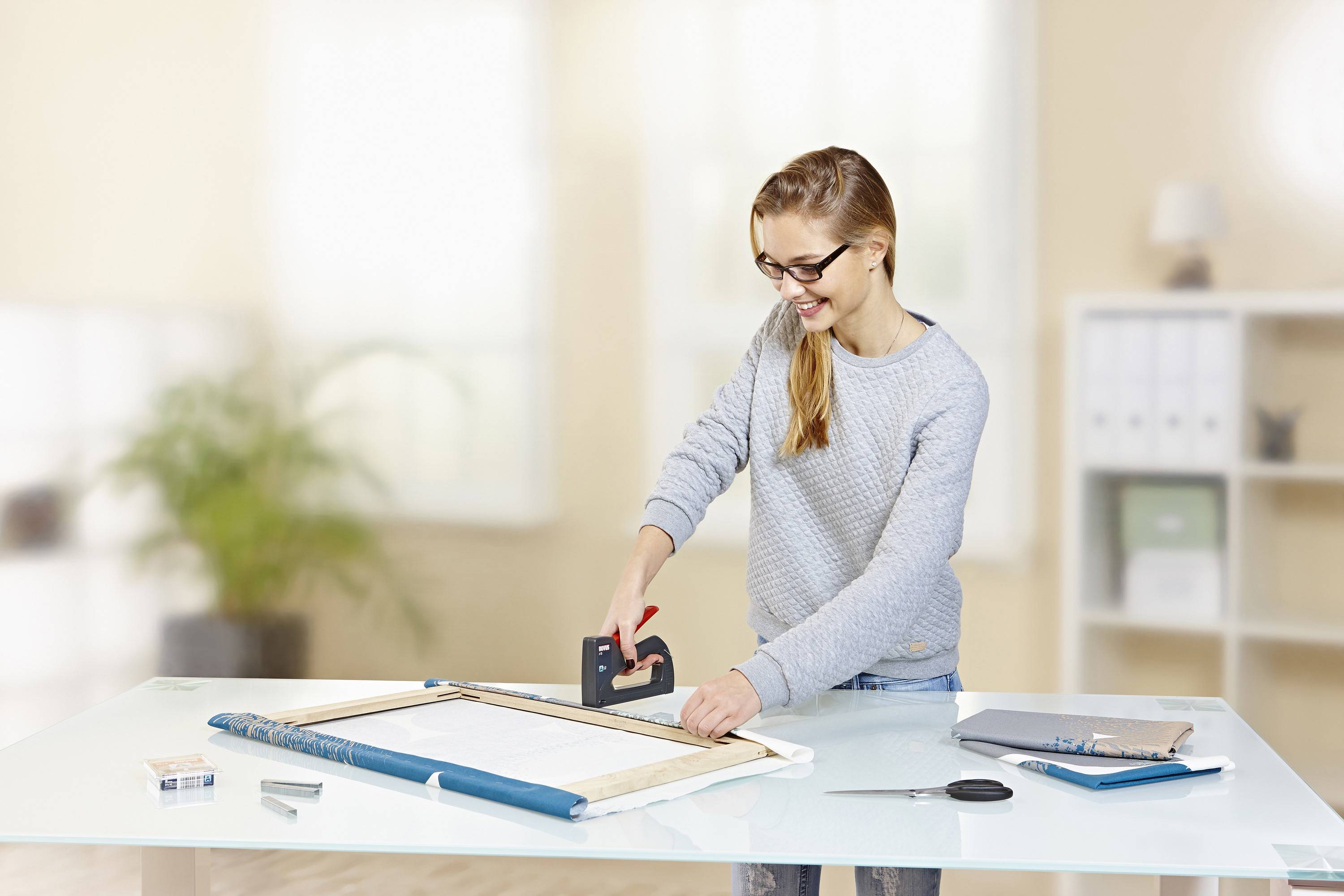 Frau mit Brille befestigt Stoff auf einem Holzrahmen mit einem Tacker in einem hellen Büro mit Tisch.
