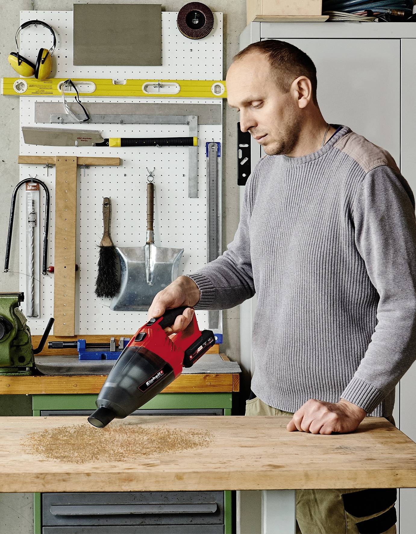 Ein Mann benutzt einen roten Handstaubsauger, um Sägespäne auf einer Werkbank in einer Werkstatt aufzuräumen. Im Hintergrund sind Werkzeuge an der Wand.