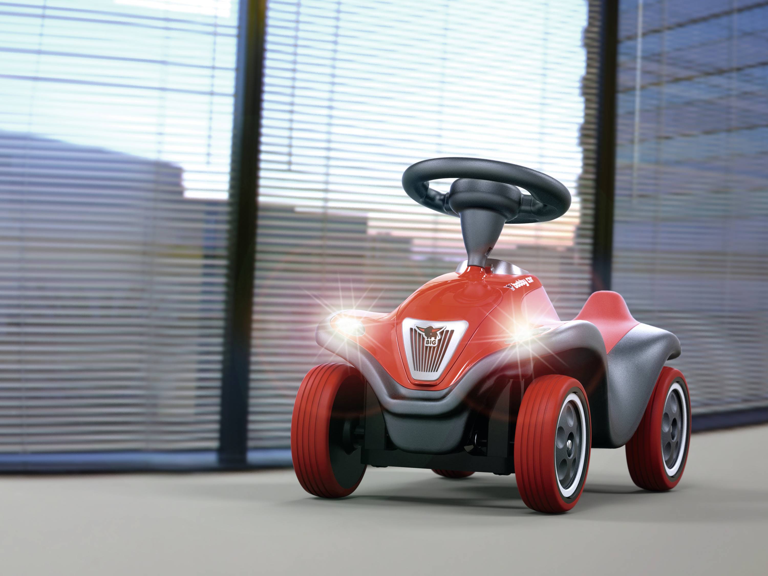 Rotes Spielzeugauto mit großen Rädern und leuchtenden Frontlichtern steht vor einem Fenster.