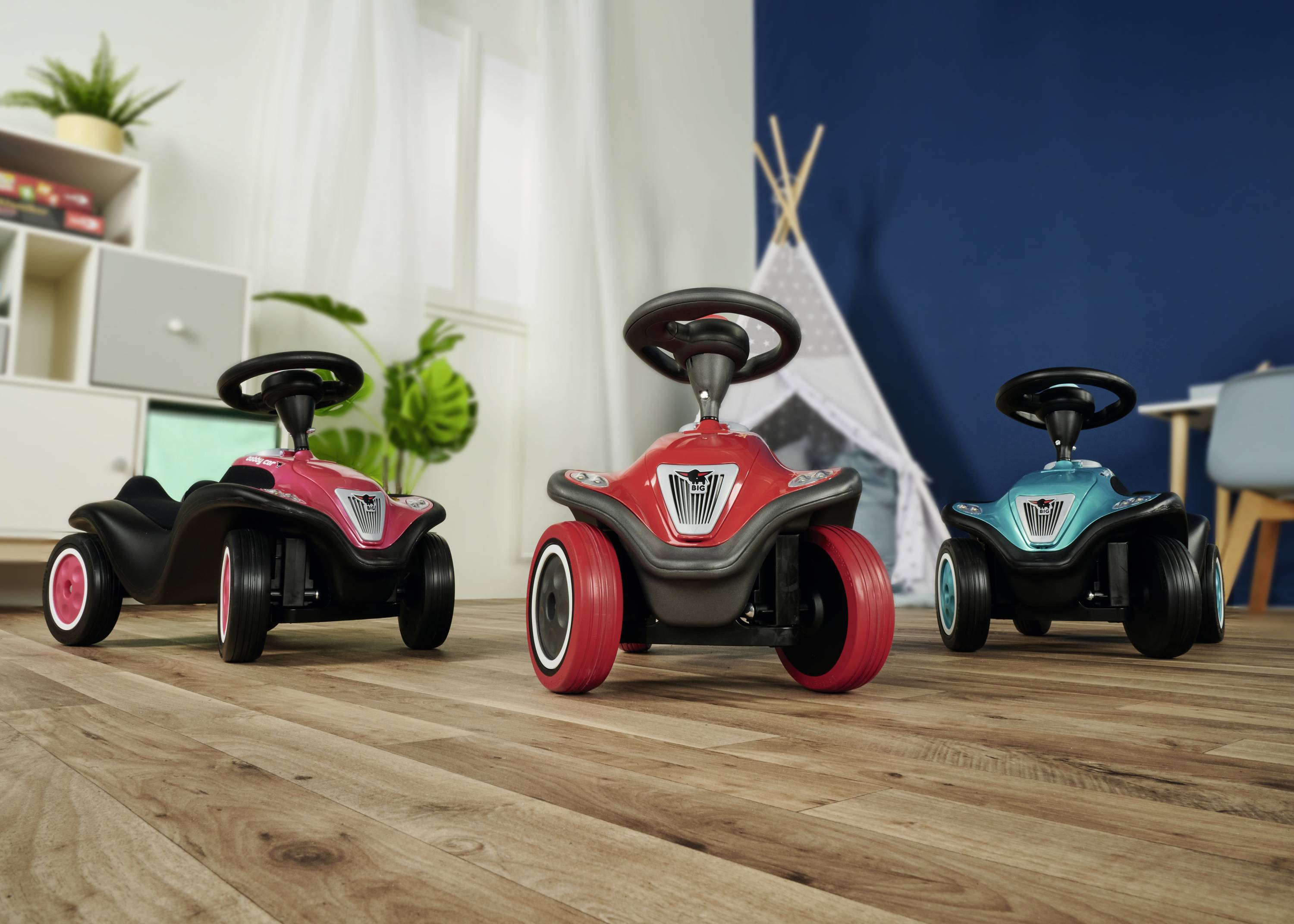 Drei farbige Rutschfahrzeuge für Kinder stehen auf einem Holzboden in einem Zimmer mit Spielzelt und Pflanzen im Hintergrund.