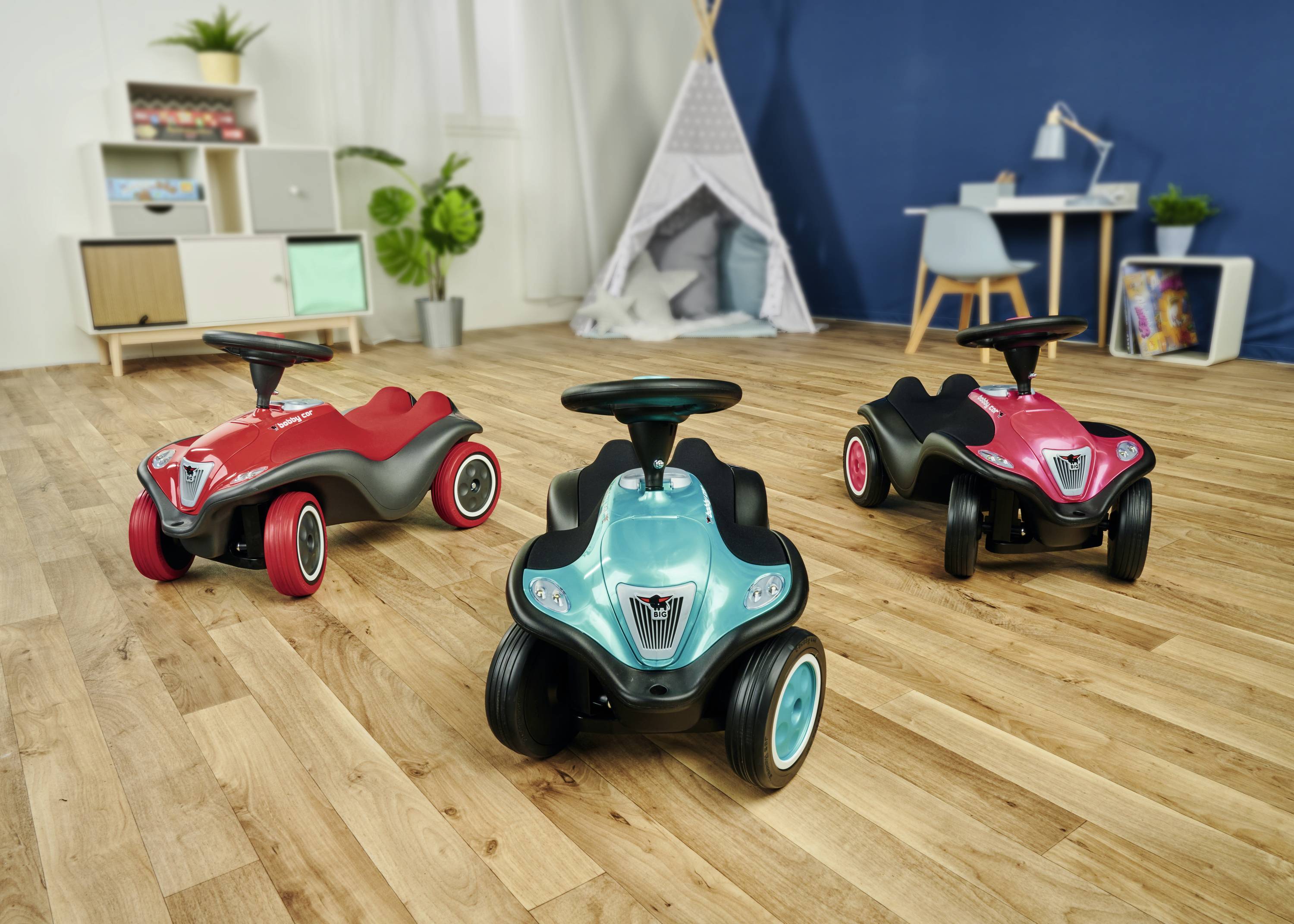 Drei bunte Kinderautos stehen in einem Raum mit Holzfußboden. Im Hintergrund befinden sich ein Regal, Pflanzen und ein Tisch mit Stuhl.