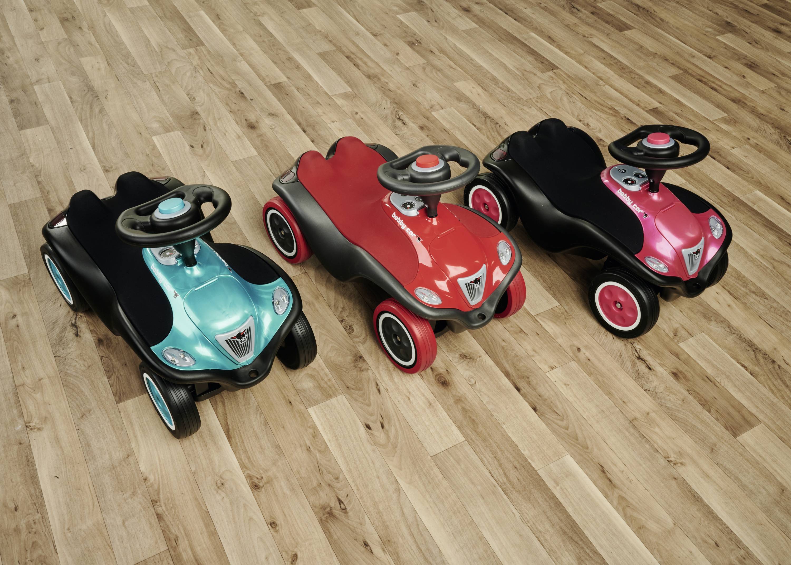 Drei Kinder-Rutschautos in Grün, Rot und Pink nebeneinander auf einem Holzfußboden, mit Lenkrädern und schwarzen Sitzen.