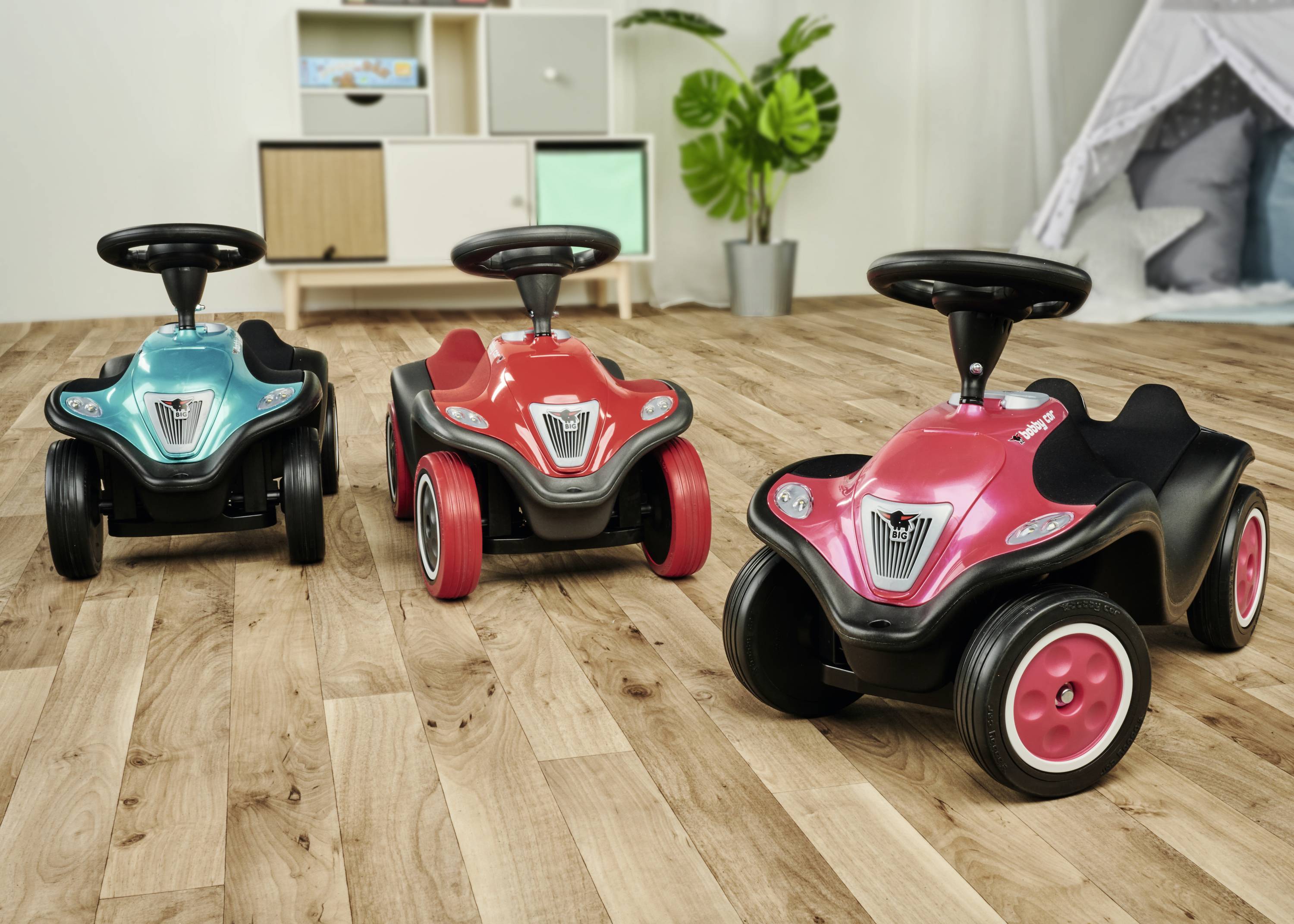 Drei bunte Kinderautos mit Lenkrädern stehen auf einem Holzfußboden. Im Hintergrund sind Regale und Pflanzen sichtbar.