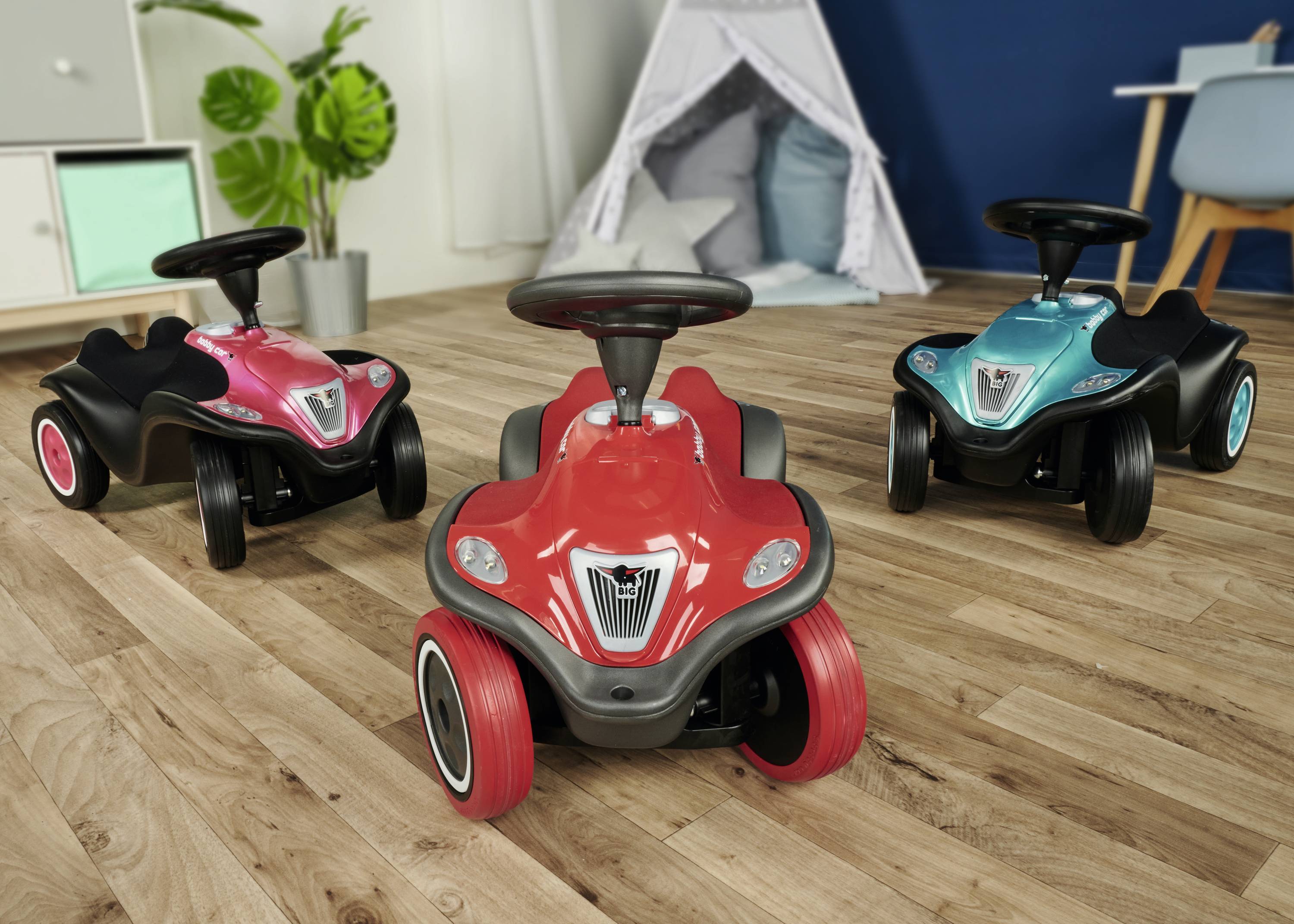 Drei bunte Rutschautos stehen auf einem Holzboden in einem Kinderzimmer. Im Hintergrund sind ein Spielzelt und ein Schreibtisch zu sehen.