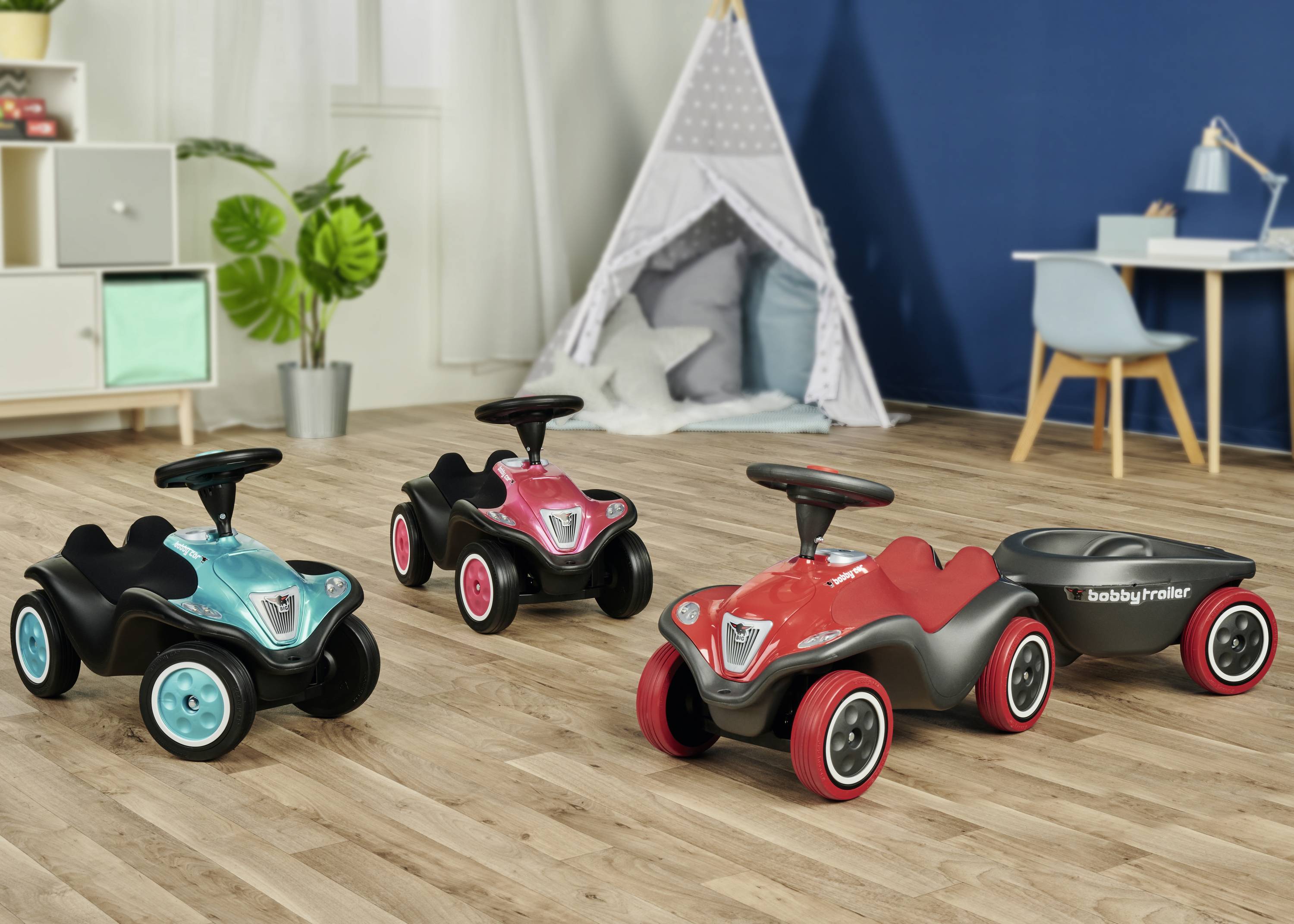 Drei Spielzeugautos in Rot, Blau und Pink stehen auf einem Holzboden in einem Kinderzimmer. Ein Anhänger ist an einem Auto befestigt.