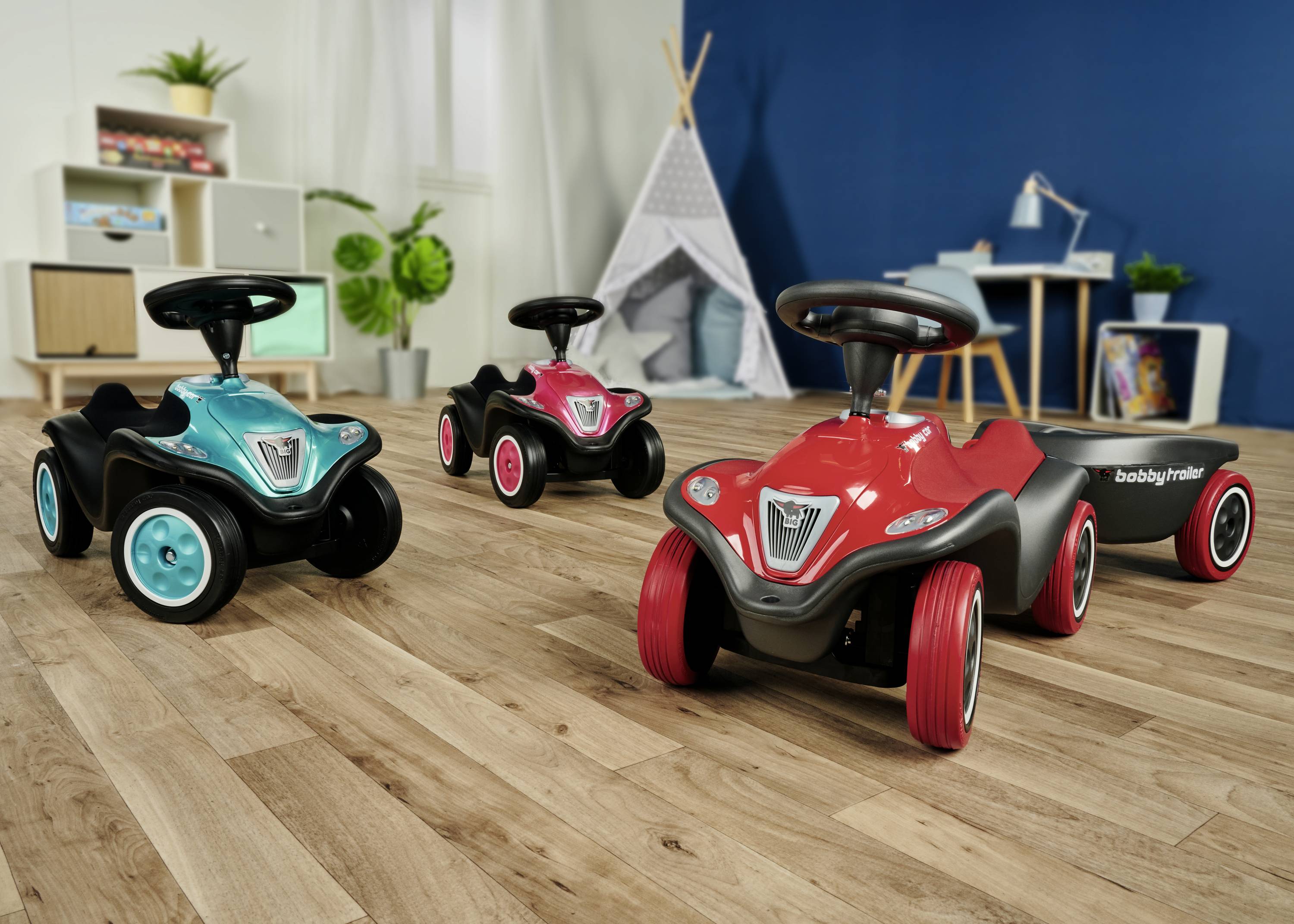 Drei Kinderfahrzeuge stehen in einem Zimmer mit Holzboden. Im Hintergrund sind ein Regal, Pflanzen und ein Kinderspielzelt sichtbar.